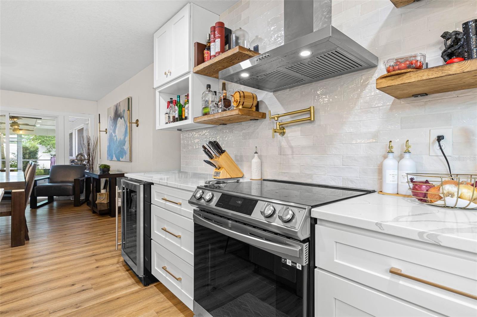A closer look at the kitchen's stylish cooking area reveals a stainless steel smooth-top range, contemporary vent hood, convenient gold pot filler, textured subway tile backsplash, and floating wood shelves. Quartz countertops and white shaker cabinetry with gold hardware complete this beautifully coordinated space.