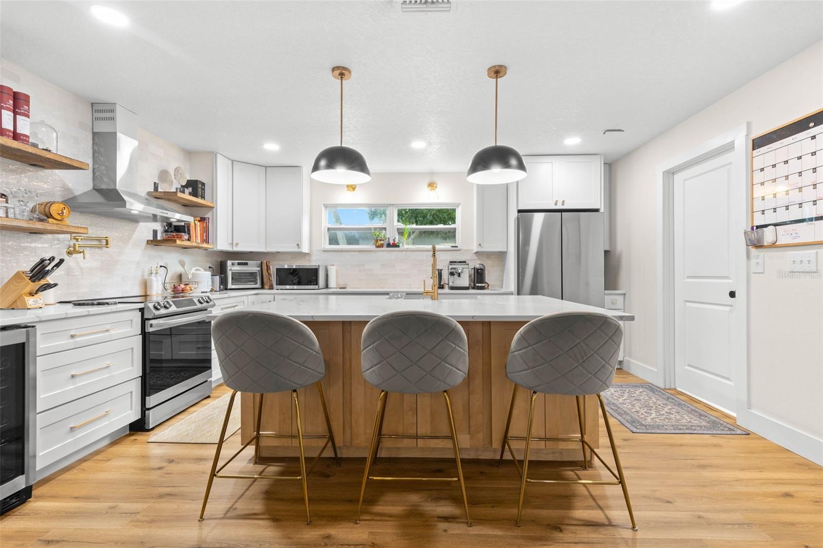 Beautifully updated and designed for both cooking and entertaining, this kitchen features an oversized quartz-topped center island with seating, white cabinetry with gold hardware, a warm wood-paneled island base, stainless steel appliances, and striking black dome pendant lights. Open shelving and a stylish tile backsplash complete the look.
