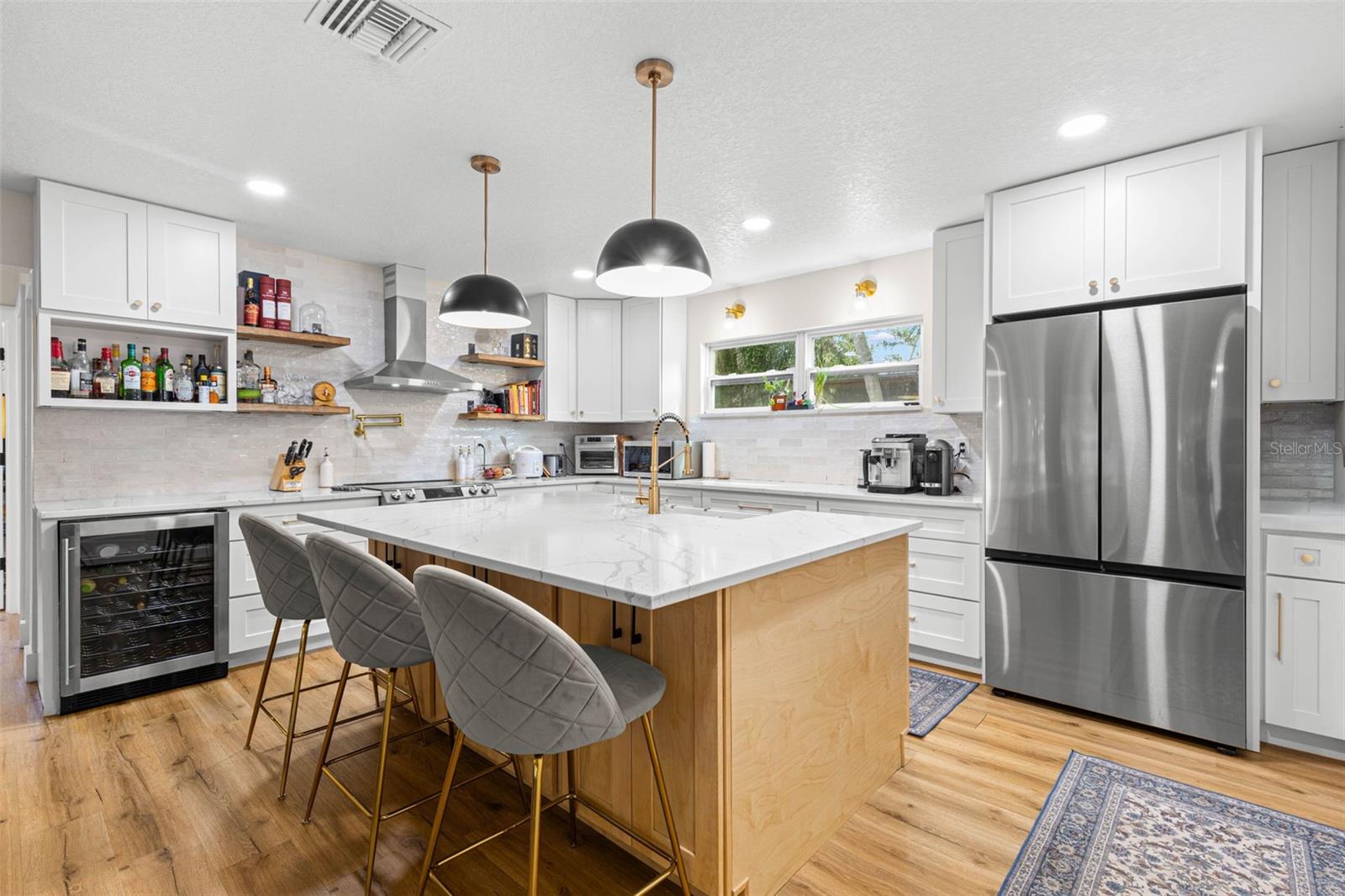 This beautifully designed kitchen combines style and practicality with abundant white shaker cabinetry, quartz countertops, stainless steel appliances, a built-in beverage refrigerator, and a generously sized center island. Floating wood shelves, gold accents, and contemporary pendant lighting add character and warmth.
