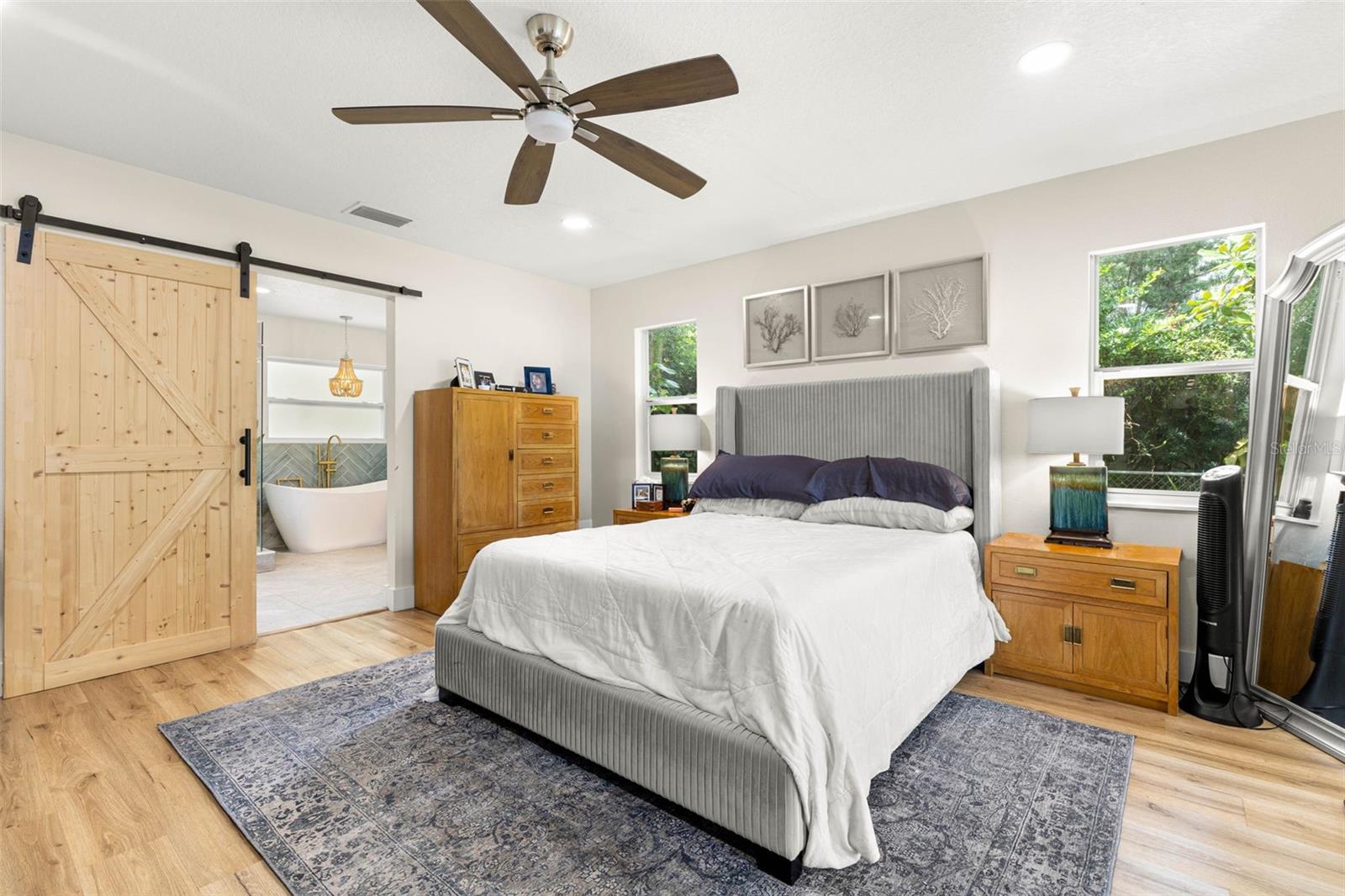 The inviting primary suite features beautiful wood-look luxury vinyl flooring, recessed lighting, a contemporary ceiling fan, and windows overlooking the greenery outside. A rustic sliding barn door adds architectural character while opening to the beautifully appointed ensuite bathroom.