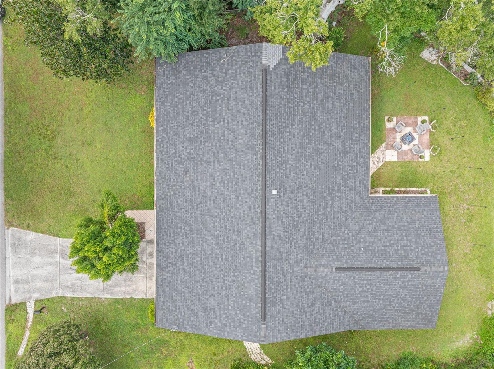 A bird-eye perspective highlights the home's expansive single-story footprint, dimensional shingle roof replaced in 2021, and surrounding lawn areas. The paver patio and fire pit area add character to the outdoor setting on this .34-acre property.