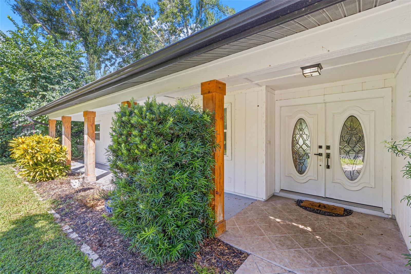 A closer look at the inviting covered front porch, featuring tiled flooring, natural wood columns, decorative double entry doors with oval glass inserts, and thoughtfully placed landscaping.