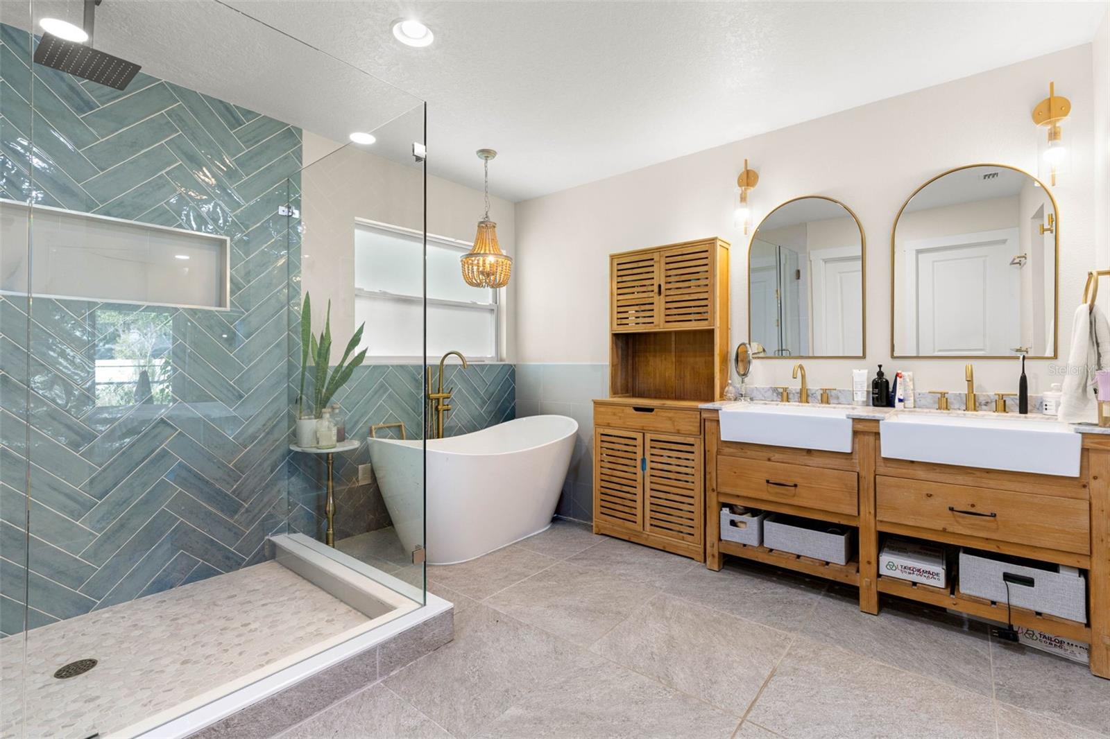 The stunning primary ensuite offers a spa-inspired retreat featuring a freestanding soaking tub, an oversized walk-in shower with frameless glass enclosure, dramatic blue-green herringbone tile, and dual farmhouse-style vessel sinks set atop warm wood vanities. Gold fixtures, arched mirrors, and coordinating sconces complete this beautifully designed bathroom.