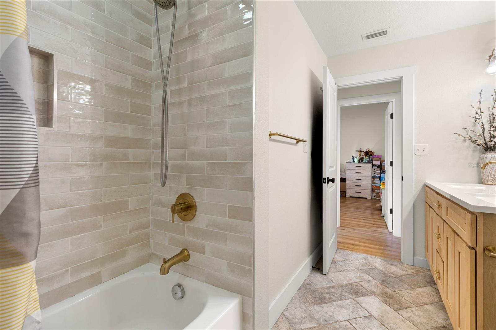 The second bathroom's tub/shower combination features glossy, neutral-toned subway tile, a recessed storage niche, and brushed gold fixtures, including a convenient handheld showerhead. Coordinating tile flooring and warm wood cabinetry complete the updated space.