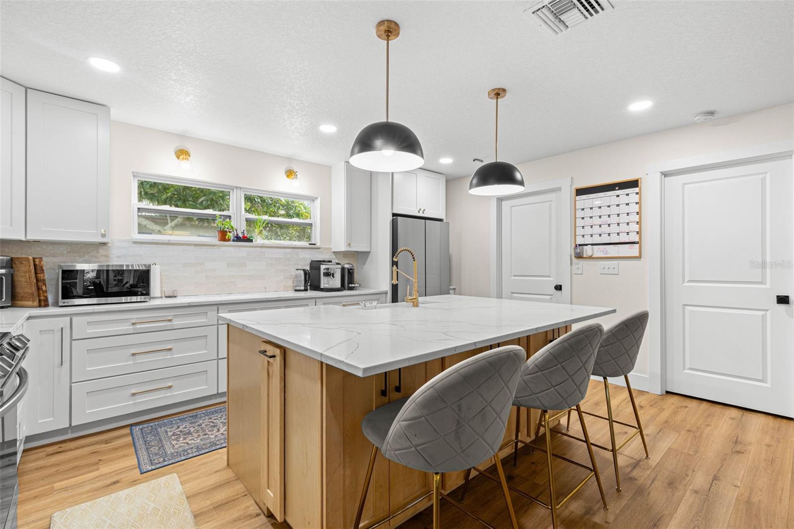 The oversized center island provides an expansive quartz countertop for meal preparation, casual dining, and entertaining. A gold gooseneck faucet, contemporary pendant lighting, abundant cabinetry, and a wide window above the counter add both style and functionality.