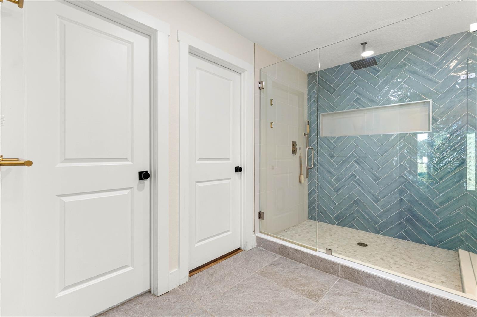 The primary bathroom's oversized walk-in shower features a frameless glass enclosure, striking blue-green herringbone tile, a rainfall showerhead, built-in recessed niche, and a pebble-style tile floor. A beautiful combination of contemporary design and everyday functionality.