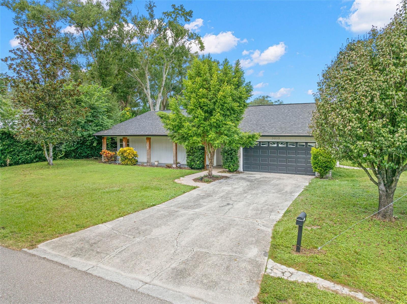 A wide concrete driveway leads to the attached two-car garage, offering convenient parking and additional storage. Mature shade trees, lush greenery, and an expansive front lawn enhance the home's curb appeal.