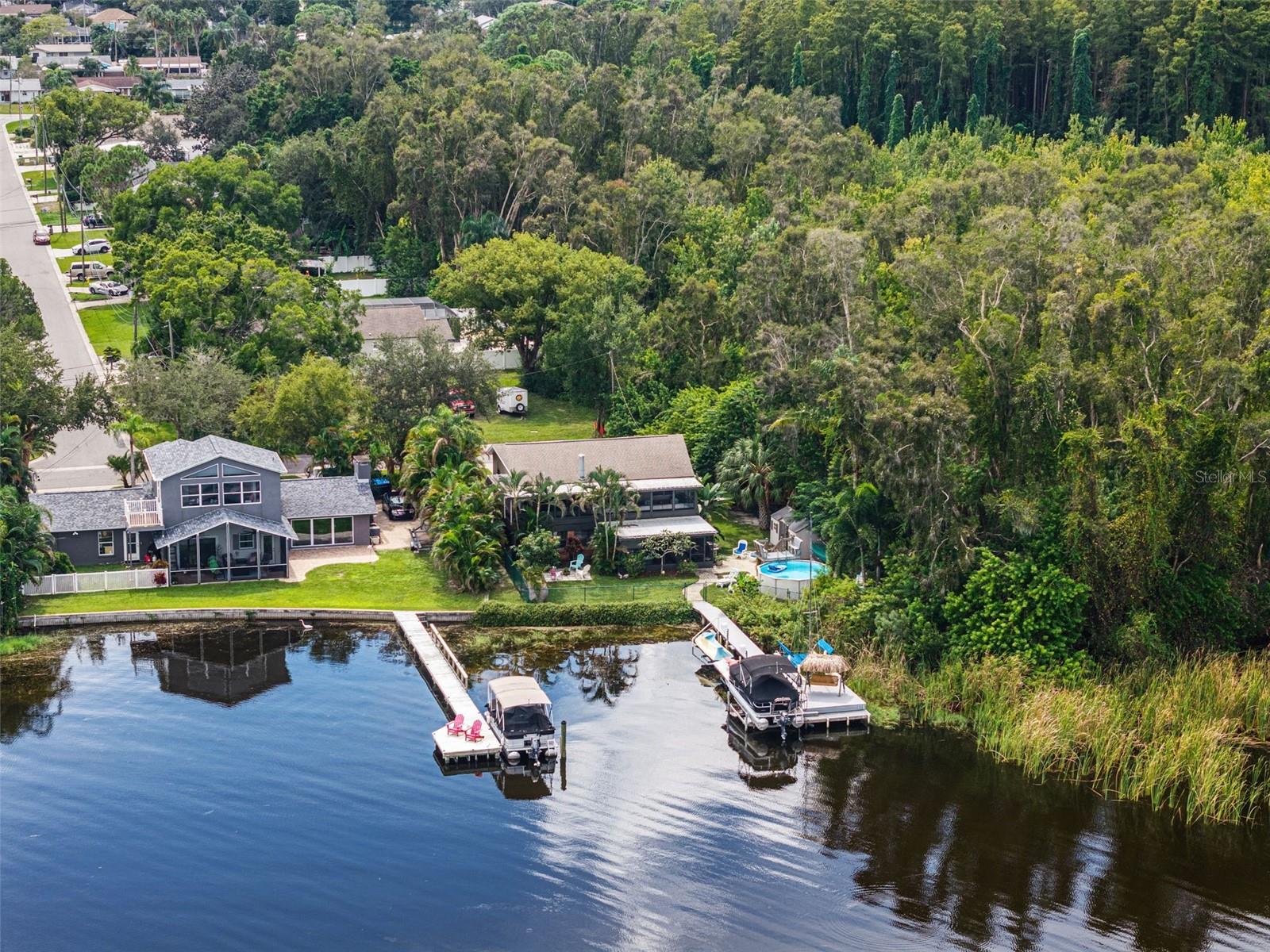 This aerial photograph brings together several of the property's standout features: an elevated home, above-ground backyard pool, private dock with pontoon boat lift, and shaded thatched-roof swing. The adjoining wooded area creates a natural backdrop, making the waterfront setting especially appealing.