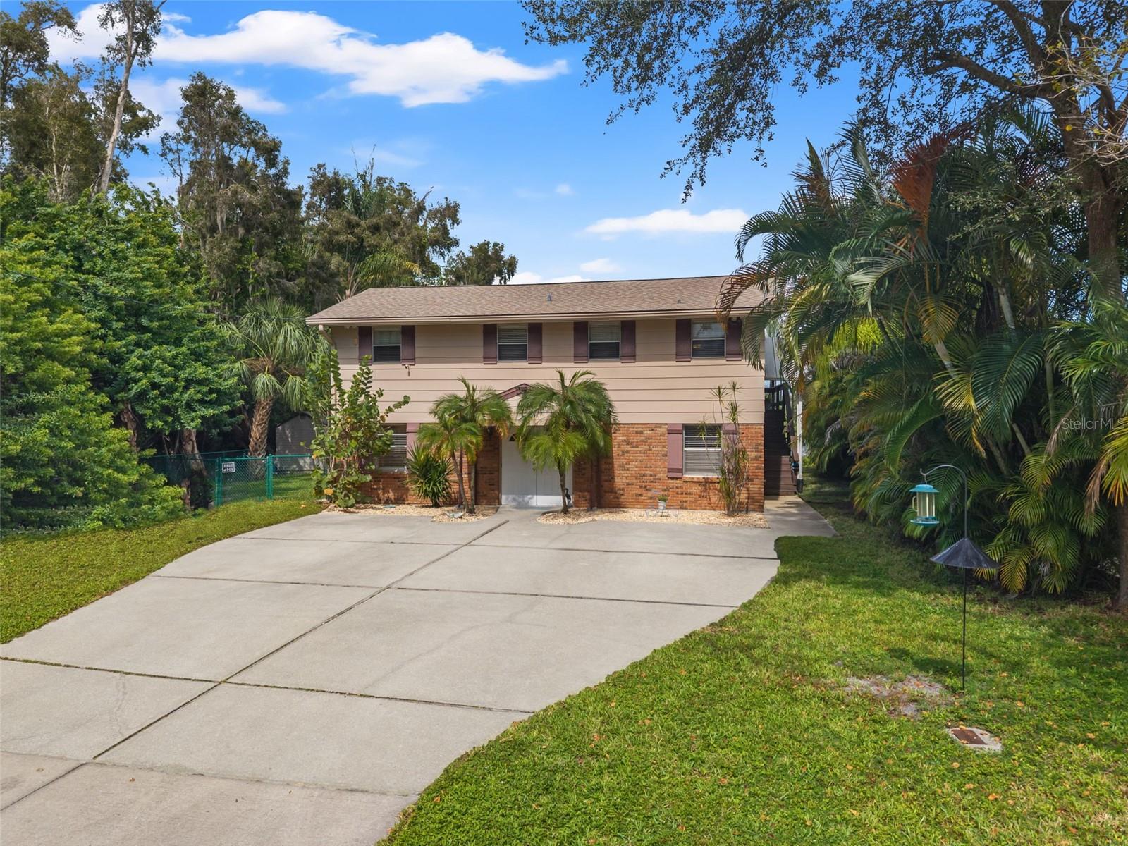Set among mature trees and tropical landscaping, this elevated home features a brick-accented lower level, a broad concrete driveway, and an upper-level living area. The spacious driveway offers generous parking, while the exterior staircase provides access to the elevated portion of the home.