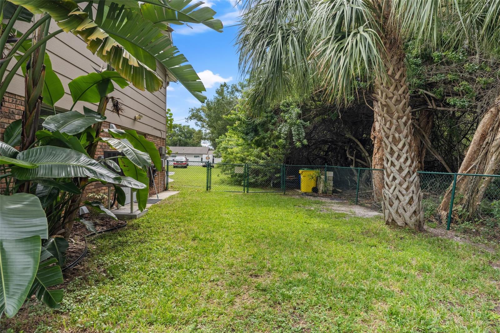 A generous grassy side yard offers additional outdoor space, framed by mature palms and lush tropical foliage. The fenced area provides room for pets, gardening, or enjoying the Florida sunshine, while a gate offers convenient access toward the front of the property.