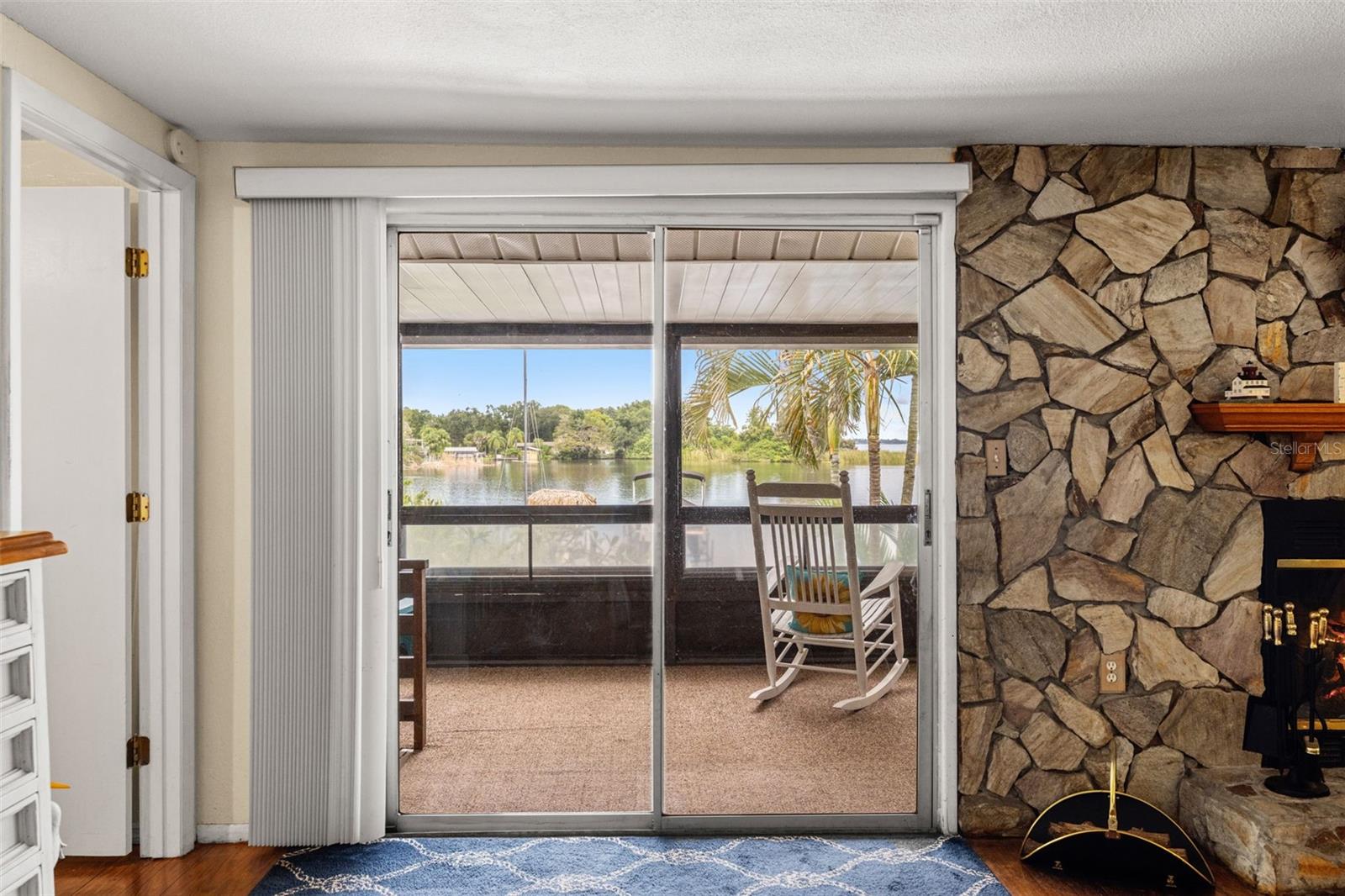 A fireplace with character! The floor-to-ceiling stone surround creates a distinctive focal point, while sliding glass doors bring in natural light and connect the living room to a screened outdoor space overlooking the water.