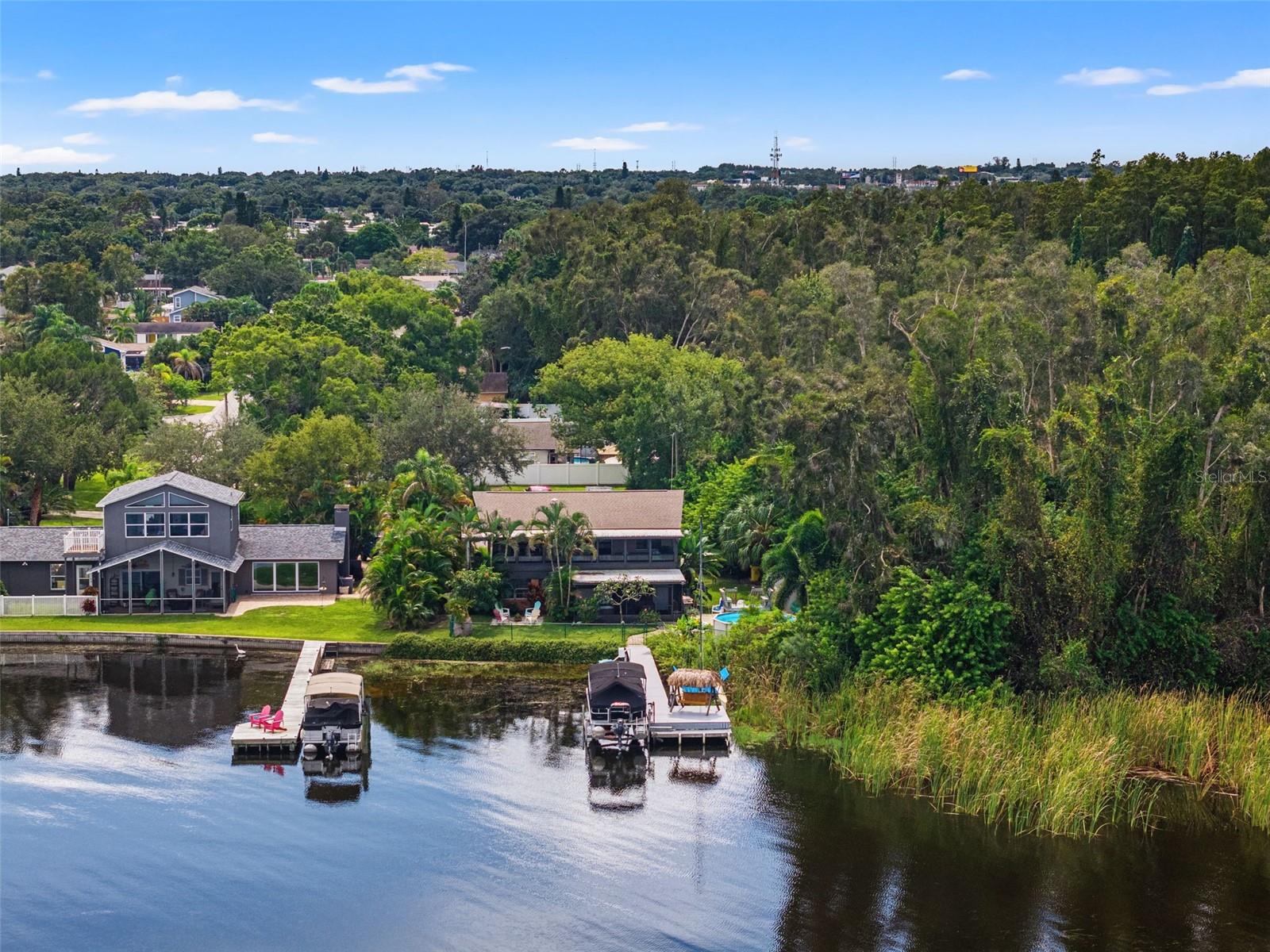 Looking toward the home from the water, this aerial photograph showcases the dock, pontoon boat lift, and shaded swing area. Tropical palms and mature trees surround the elevated residence, while natural shoreline vegetation contributes to the property's peaceful waterfront atmosphere.