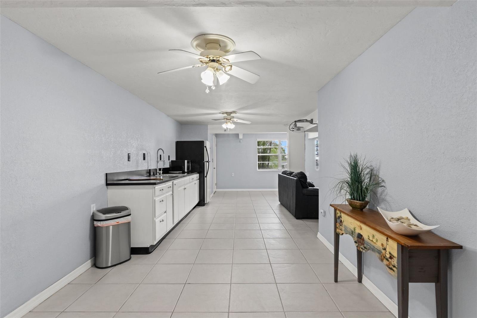 The home's lower level offers a convenient kitchenette with white cabinetry, contrasting dark countertops, a sink, refrigerator, and microwave. Neutral tile flooring extends throughout the space, connecting the kitchenette with the adjoining living area. A practical setup for entertaining, hosting guests, or enjoying additional space separate from the main living level.