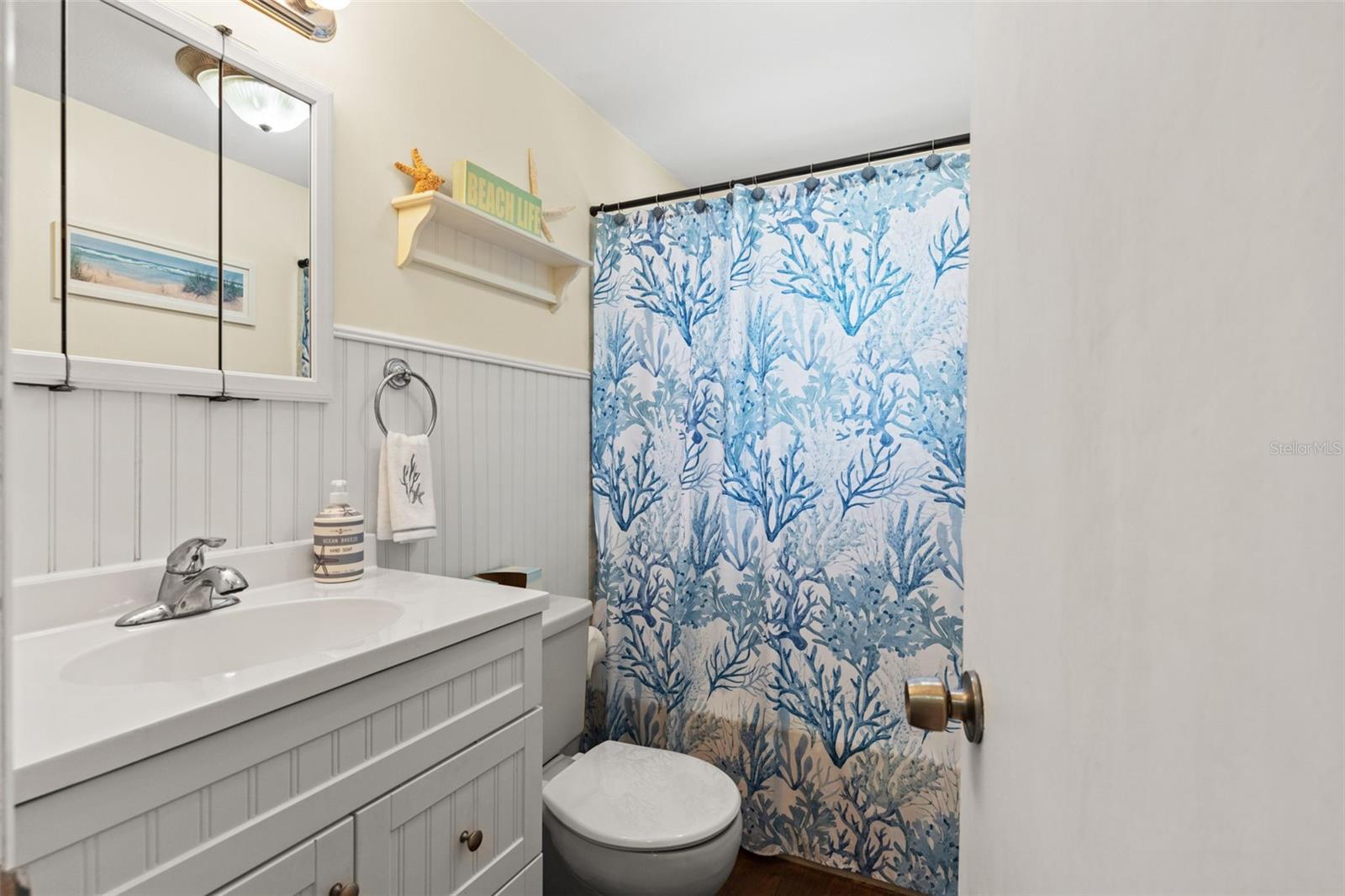 Coastal charm continues into this full bathroom, featuring a white vanity with decorative beadboard-style detailing, a mirrored medicine cabinet, and a tub/shower combination. White wainscoting and cheerful blue accents complete the beach-inspired look.
