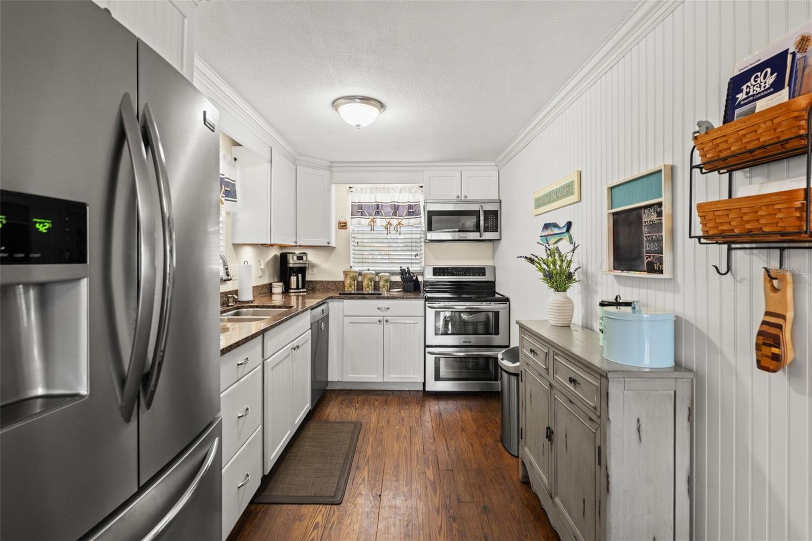 This inviting kitchen combines white cabinetry, stainless steel appliances, wood flooring, and warm-toned countertops. Crown molding and beadboard-style wall detailing add character, while the galley layout keeps everything conveniently within reach.