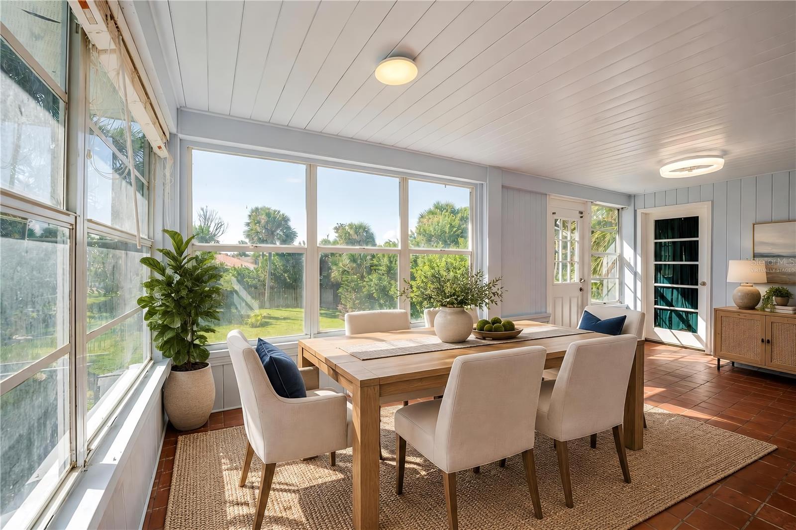 Virtually Staged - Breakfast nook featuring red tile floors, light-colored wood paneling, flush-mount ceiling lights, and wide windows overlooking the green backyard. Exterior glass doors provide easy access to the lawn and surrounding grounds.