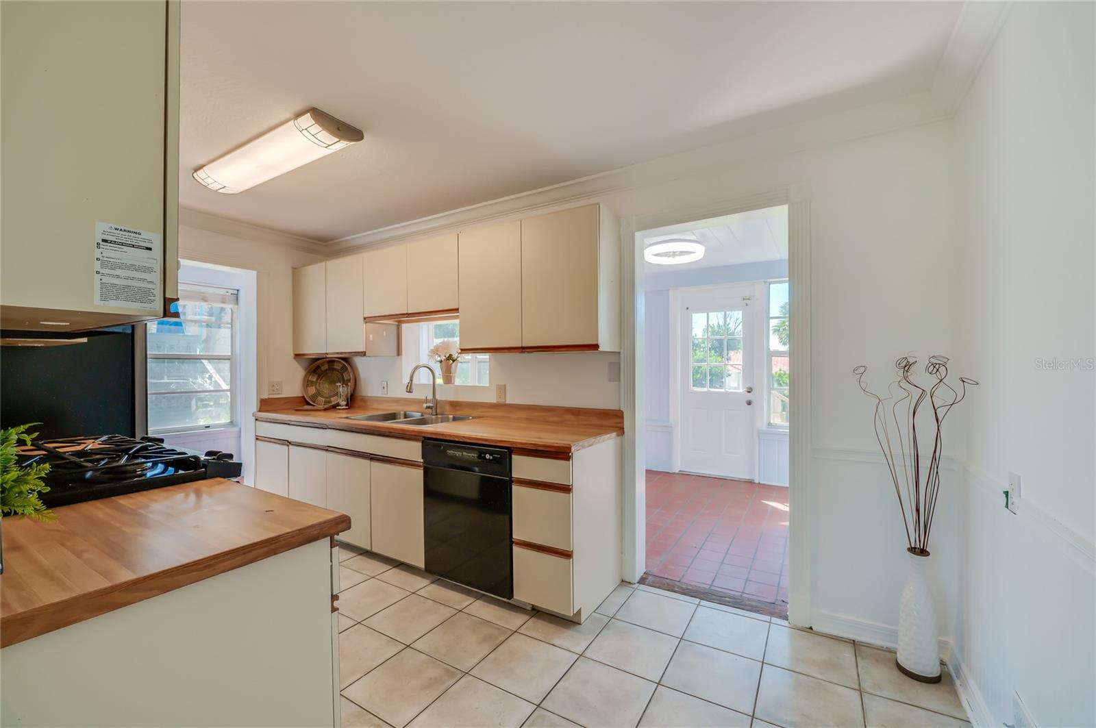 Functional kitchen featuring butcher block-style countertops, classic cabinetry, tile flooring, and integrated black appliances. An adjacent open doorway provides direct access to a sunlit room with terracotta tile floors.