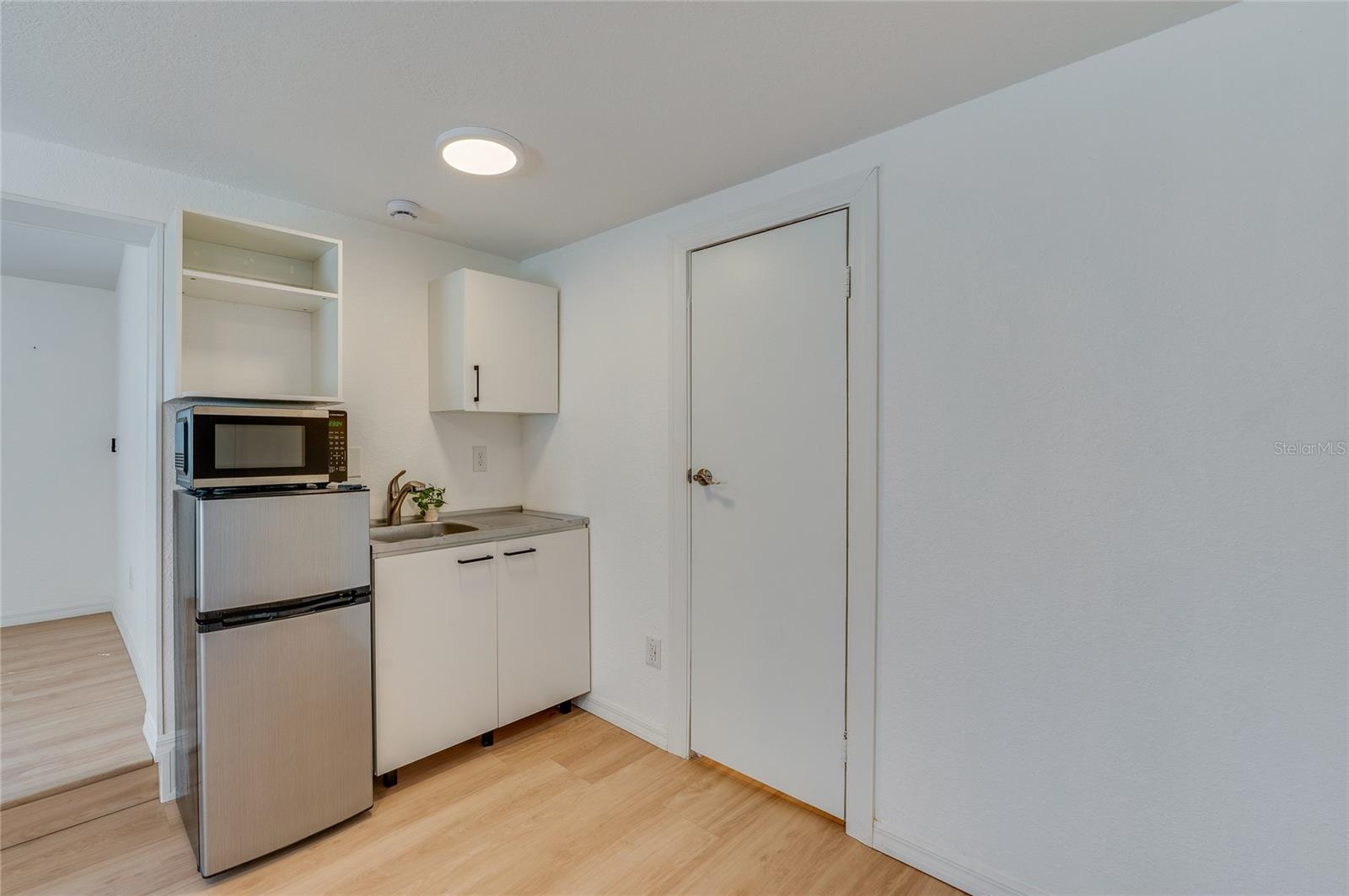 Functional kitchenette area equipped with a compact stainless-steel mini refrigerator, microwave, and white cabinetry with a sink. Clean white walls and warm LVP floors complete the modern, low-maintenance design.