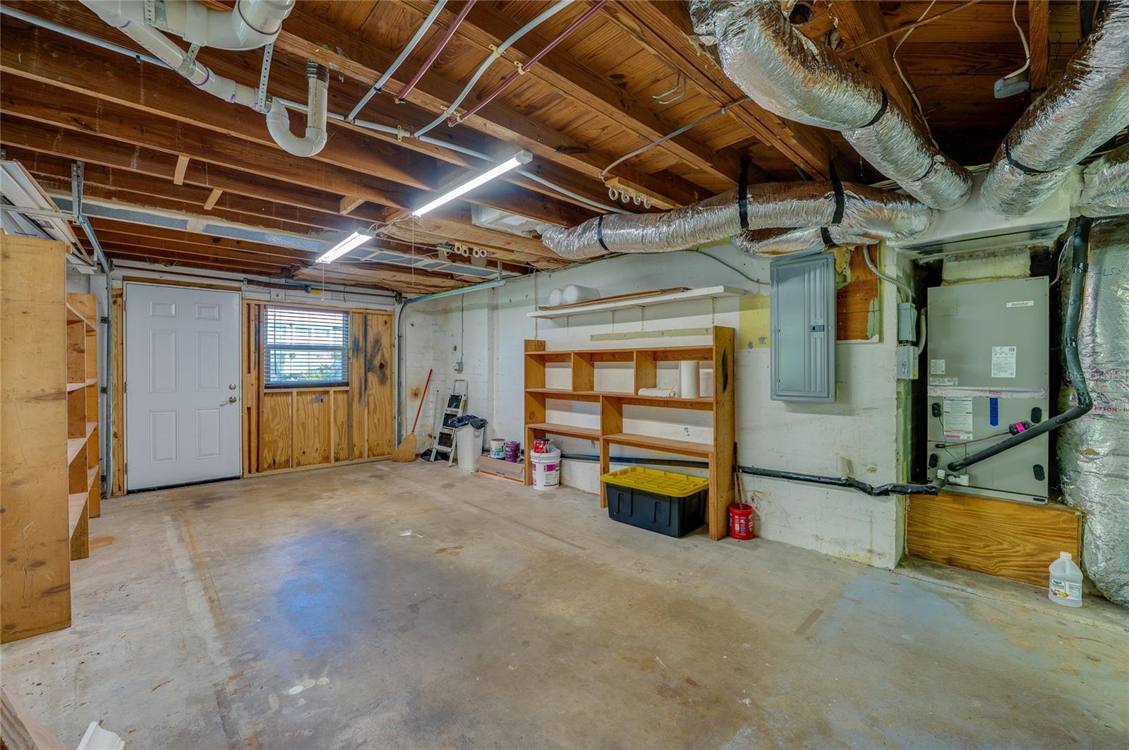 Functional utility room showcasing exposed overhead piping, ductwork, an HVAC unit, and an electrical panel. Built-in wooden shelving and a concrete slab floor offer practical storage space.