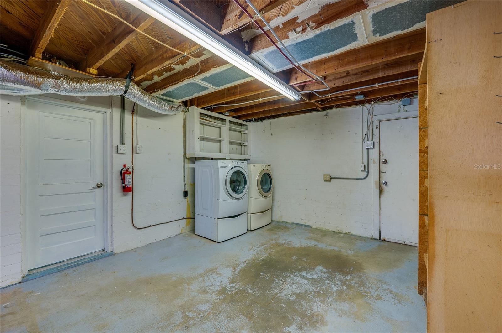 Dedicated laundry area equipped with a side-by-side washer and dryer set underneath overhead wall cabinets. The space features durable concrete floors, exposed ceiling beams, and direct door access.