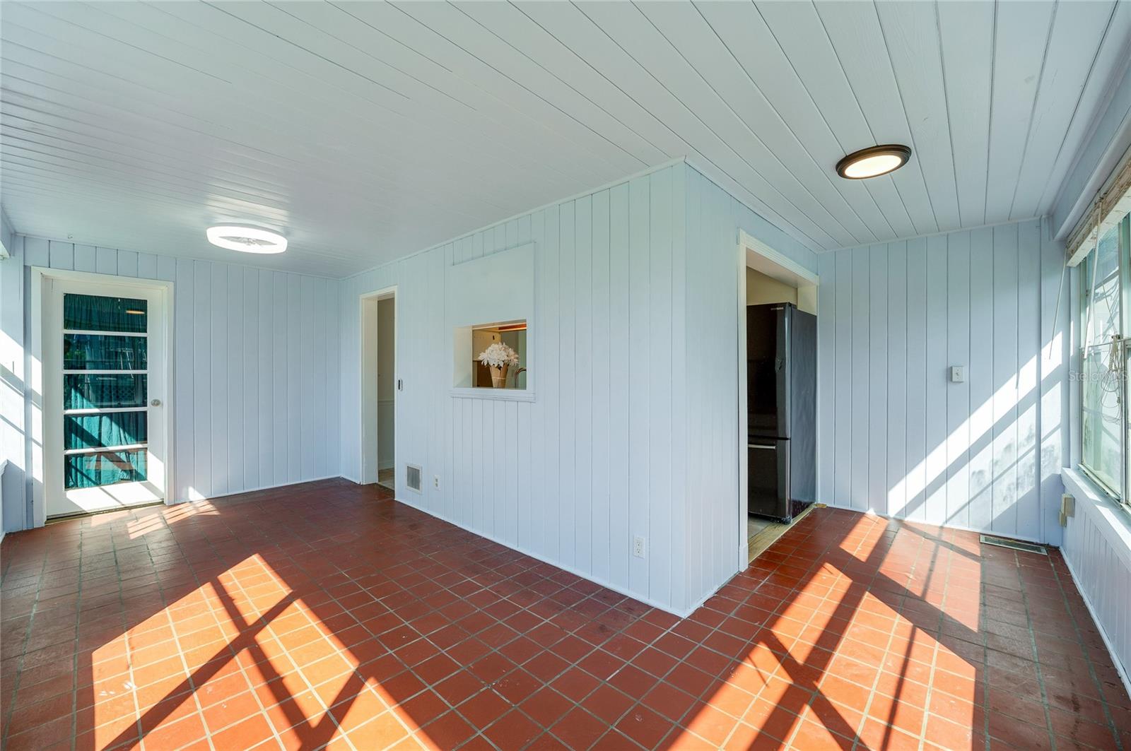 Sun-filled dining area and breakfast nook showcasing red tile flooring, modern flush-mount lighting, and white paneled walls. Large windows fill the space with natural light while direct door access leads outdoors.