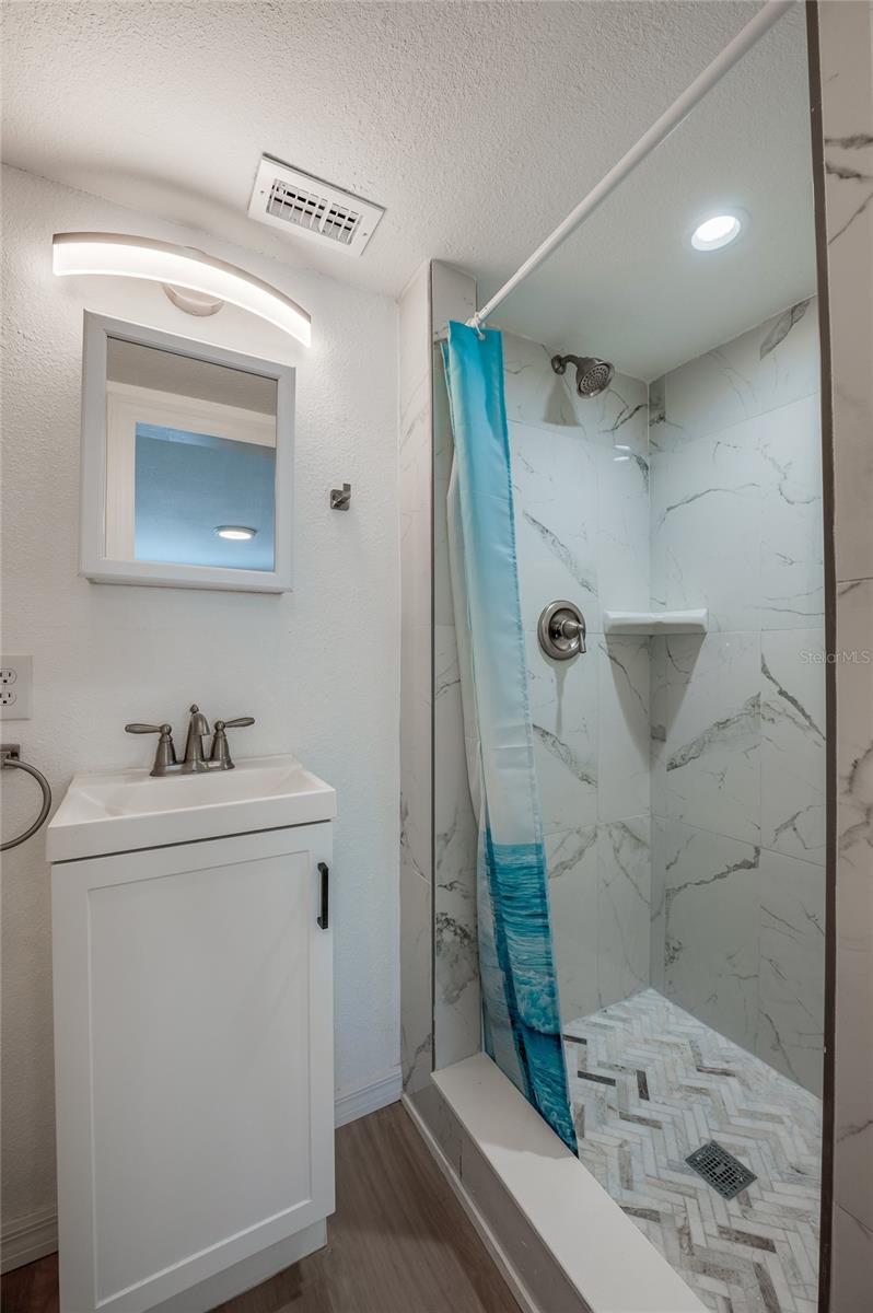 Modernized bathroom featuring a white single-sink vanity with modern hardware and a wall-mounted mirror. The walk-in shower is styled with marble-patterned wall tile and intricate herringbone floor tiling.