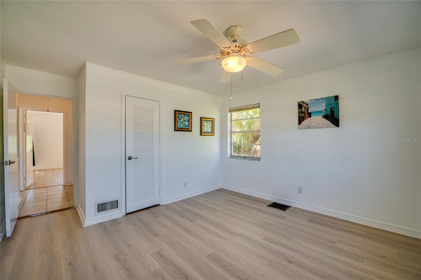 Clean interior space showcasing light flooring, a white ceiling fan, and a louvered closet door. An open doorway leads out into a tiled hallway connecting to the rest of the layout.