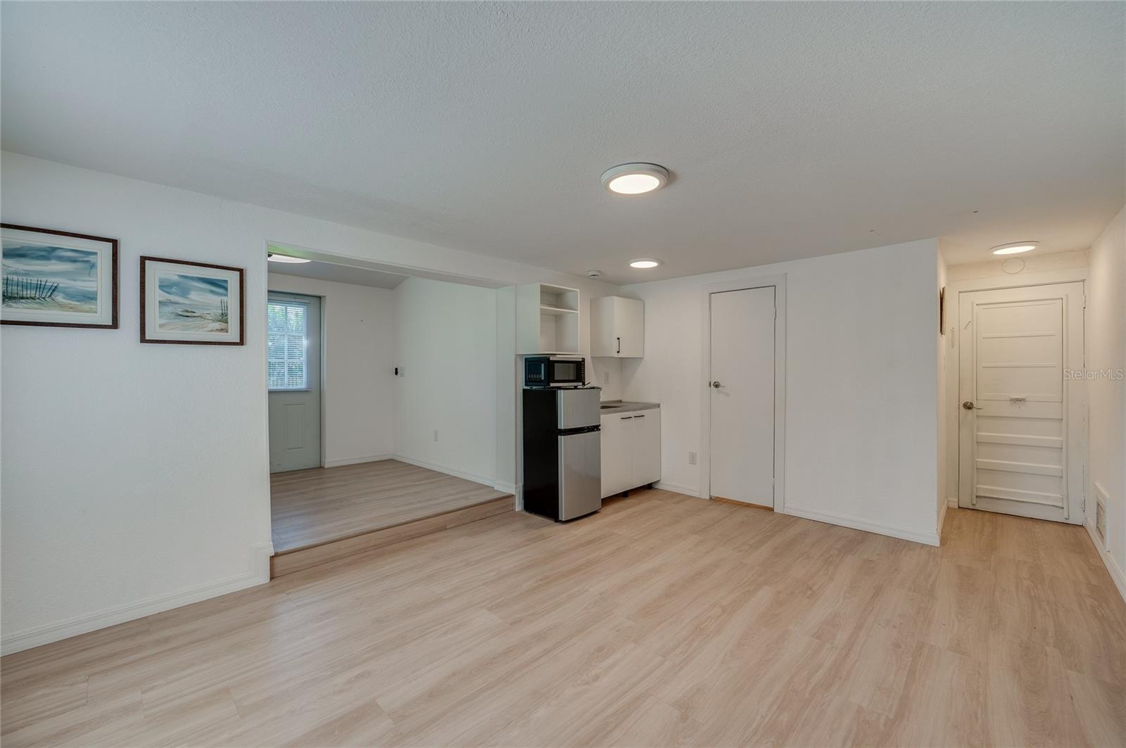 Spacious open-concept living area featuring light LVP flooring, recessed ceiling lighting, and a compact kitchenette nook. An adjacent step-up alcove provides additional versatile living space.