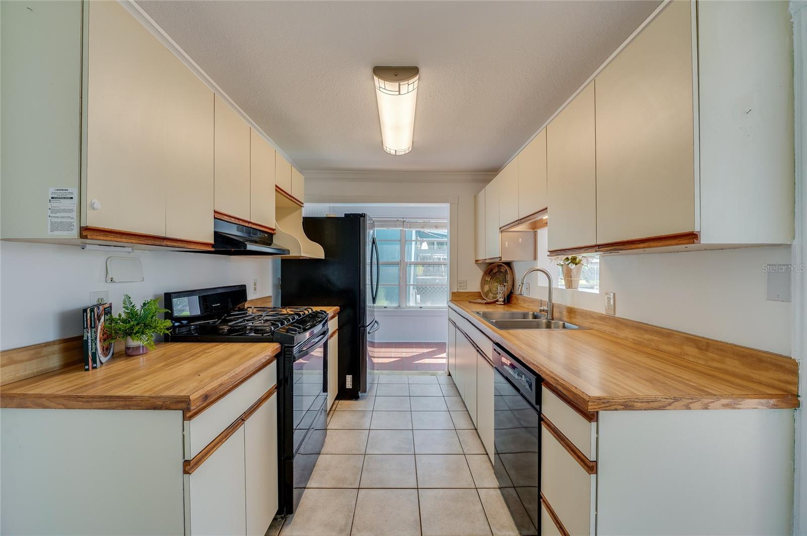 Galley-style kitchen design equipped with dual-sided cabinetry, warm wood-toned countertops, a double-basin sink, and overhead light fixtures. The walkthrough layout opens up directly to the sunroom beyond.