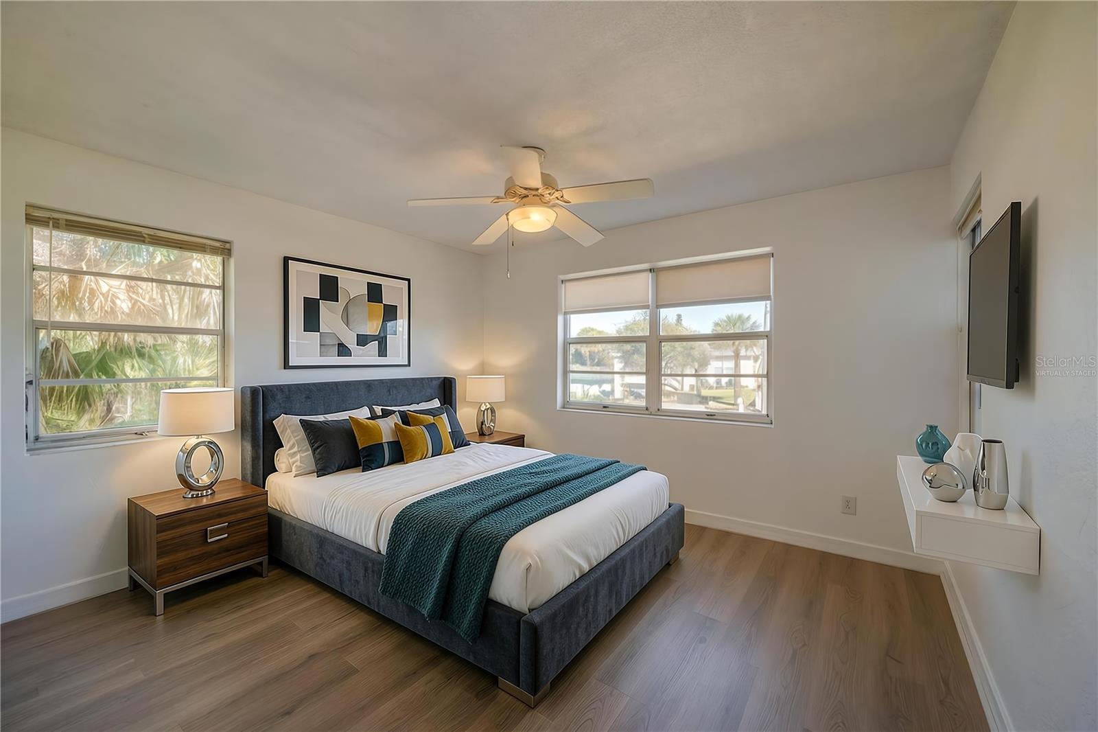 Virtually Staged - Bright bedroom area framed by continuous LVP flooring, smooth white walls, and a traditional white ceiling fan. Dual aspect windows bring in abundant sunlight and views of the surrounding palm trees.
