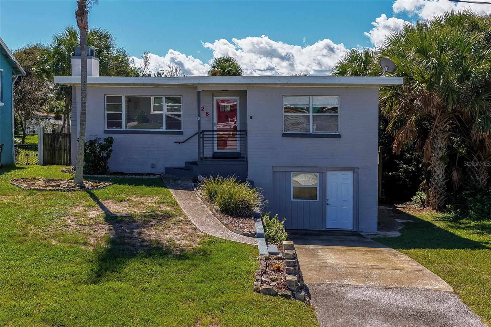 Exterior view of a coastal single-family home featuring a front driveway, grassy yard, and surrounding palm trees. The light gray facade offers elevated main entry steps and a lower ground-level entryway. Exterior view of a coastal single-family home featuring a front driveway, grassy yard, and surrounding palm trees. The light gray facade offers elevated main entry steps and a lower ground-level entryway.