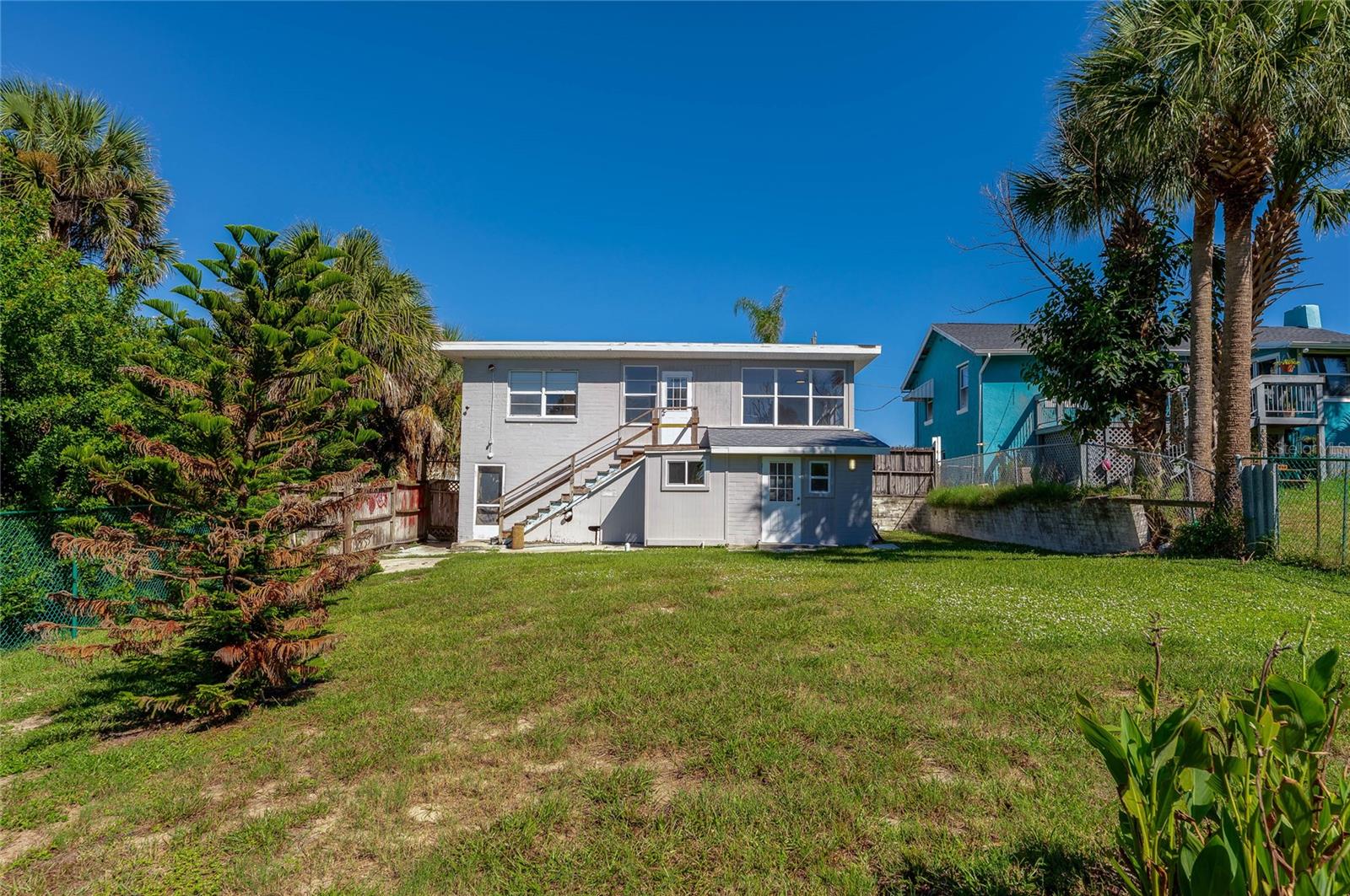 Rear view of a two-story home featuring an upper-level exterior staircase and a spacious grassy backyard. Vibrant palm trees and surrounding greenery enhance the coastal outdoor setting.