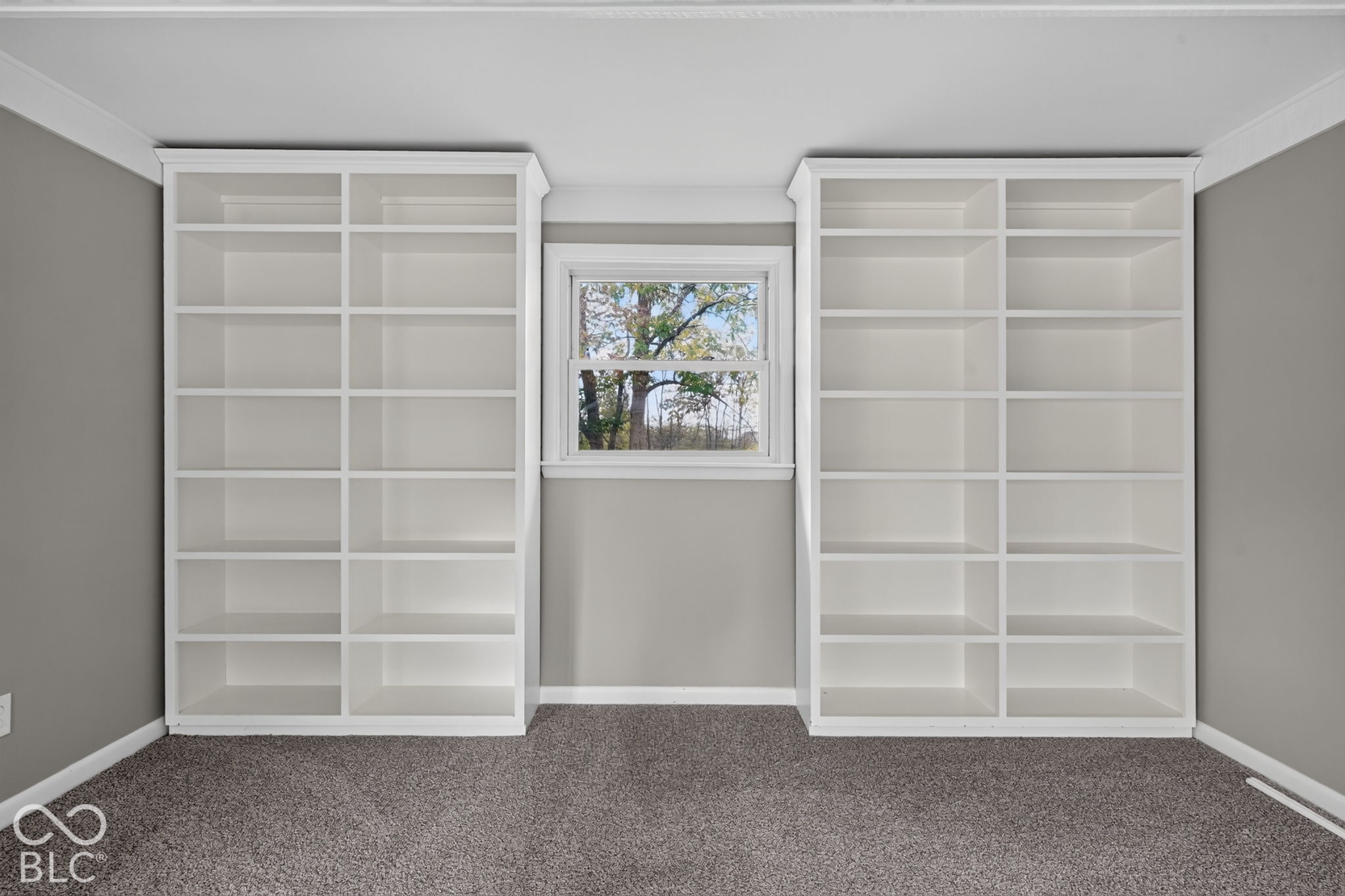 Floor-to-ceiling built-in white shelving, crown molding, and a central window with exterior tree-lined scenery
