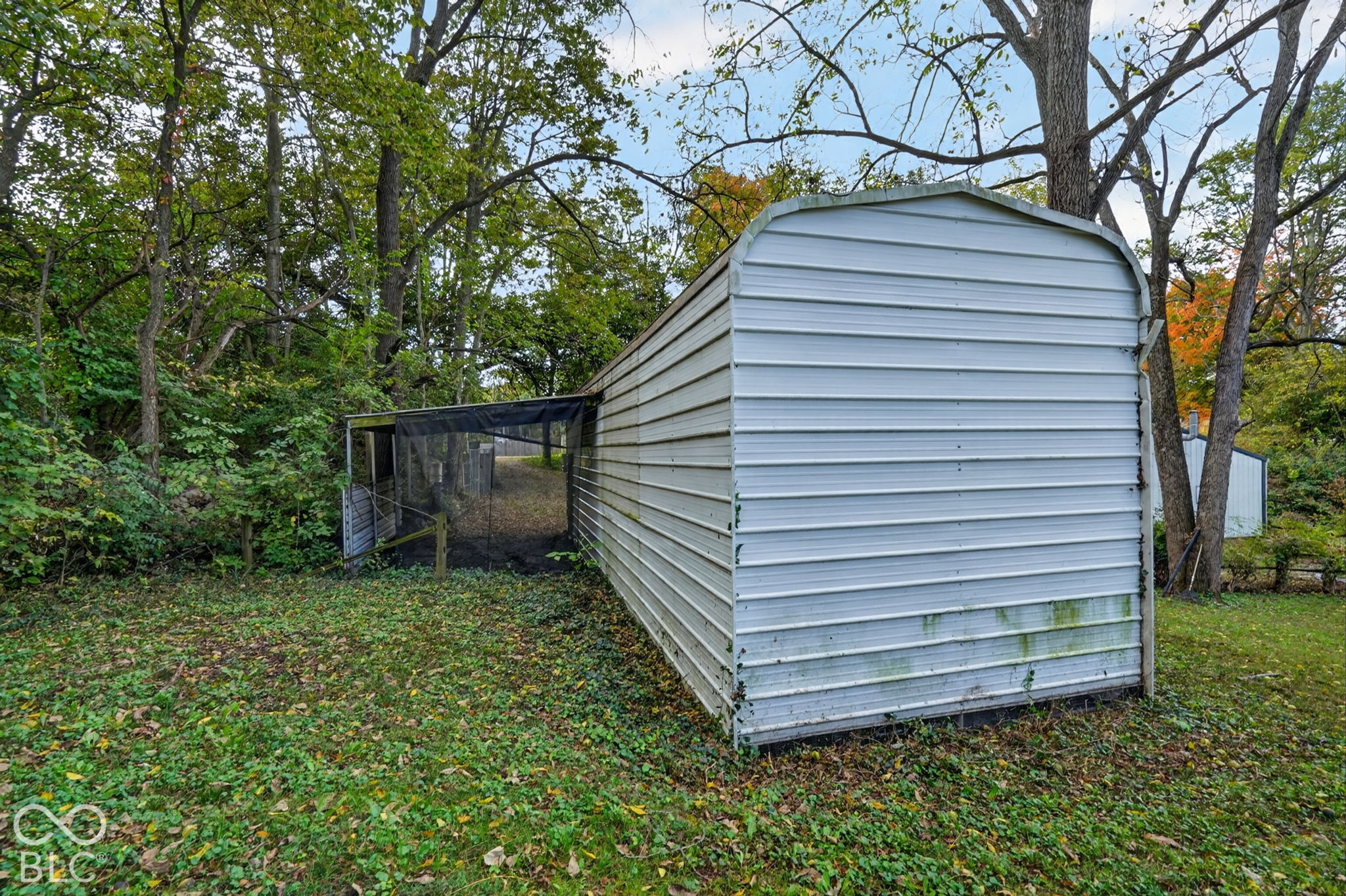 Metal storage structure featuring horizontal siding and a mesh-enclosed external area on a lot with mature trees