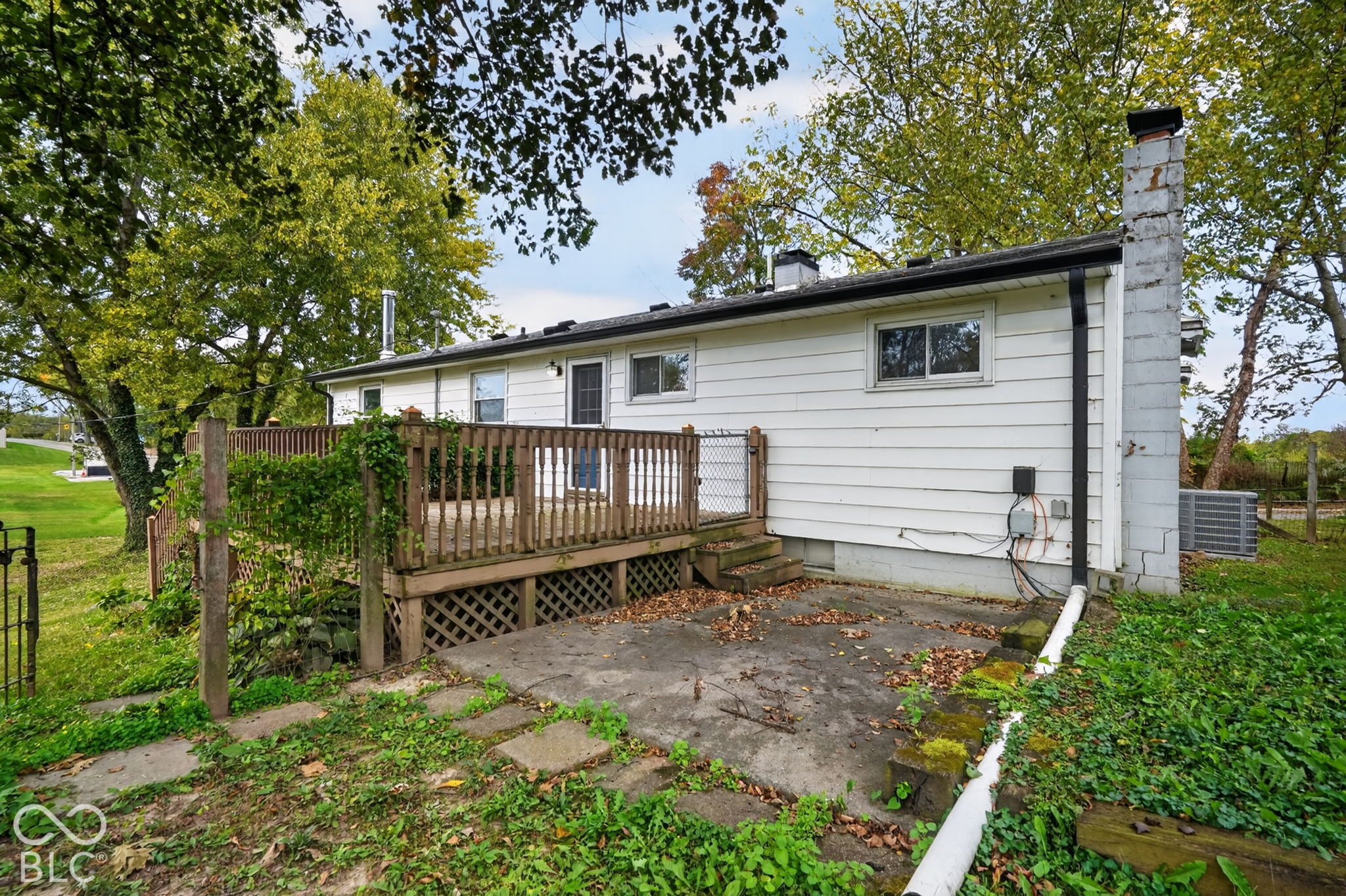 Rear exterior featuring white siding, a raised wood deck with balustrades, a ground-level concrete patio, and a masonry chimney