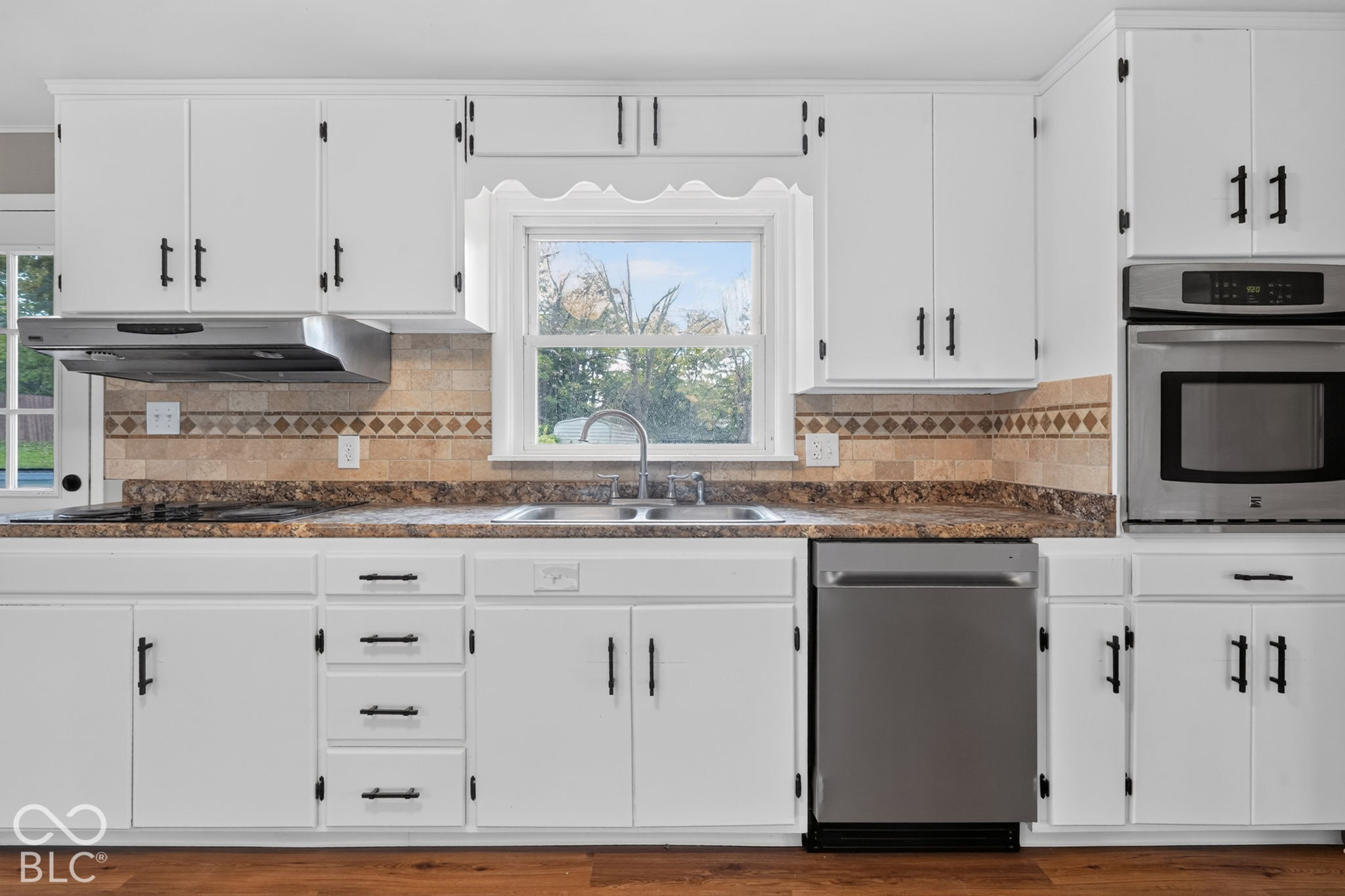 Kitchen featuring white cabinetry with matte black hardware, stone-patterned countertops, a decorative tile backsplash with border inlay, stainless steel appliances, and a double-basin stainless steel sink
