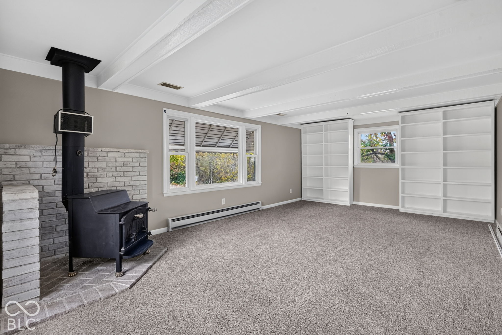 Exposed white ceiling beams, built-in floor-to-ceiling shelving, painted brick hearth, and a freestanding wood-burning stove
