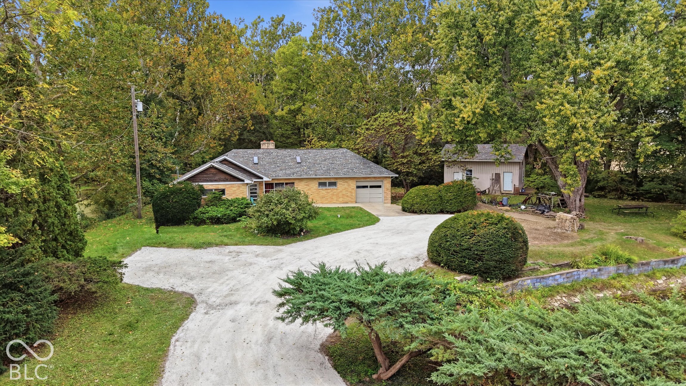 Single-story brick exterior with an attached garage, featuring a gravel driveway, mature trees, and an additional detached structure