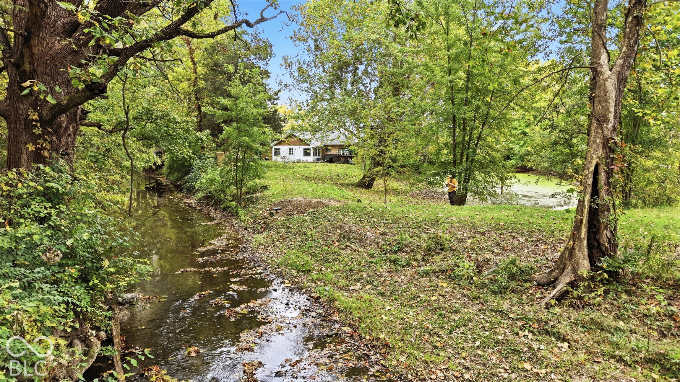 White exterior structure situated on a grassy incline, bordered by mature trees and a natural stream