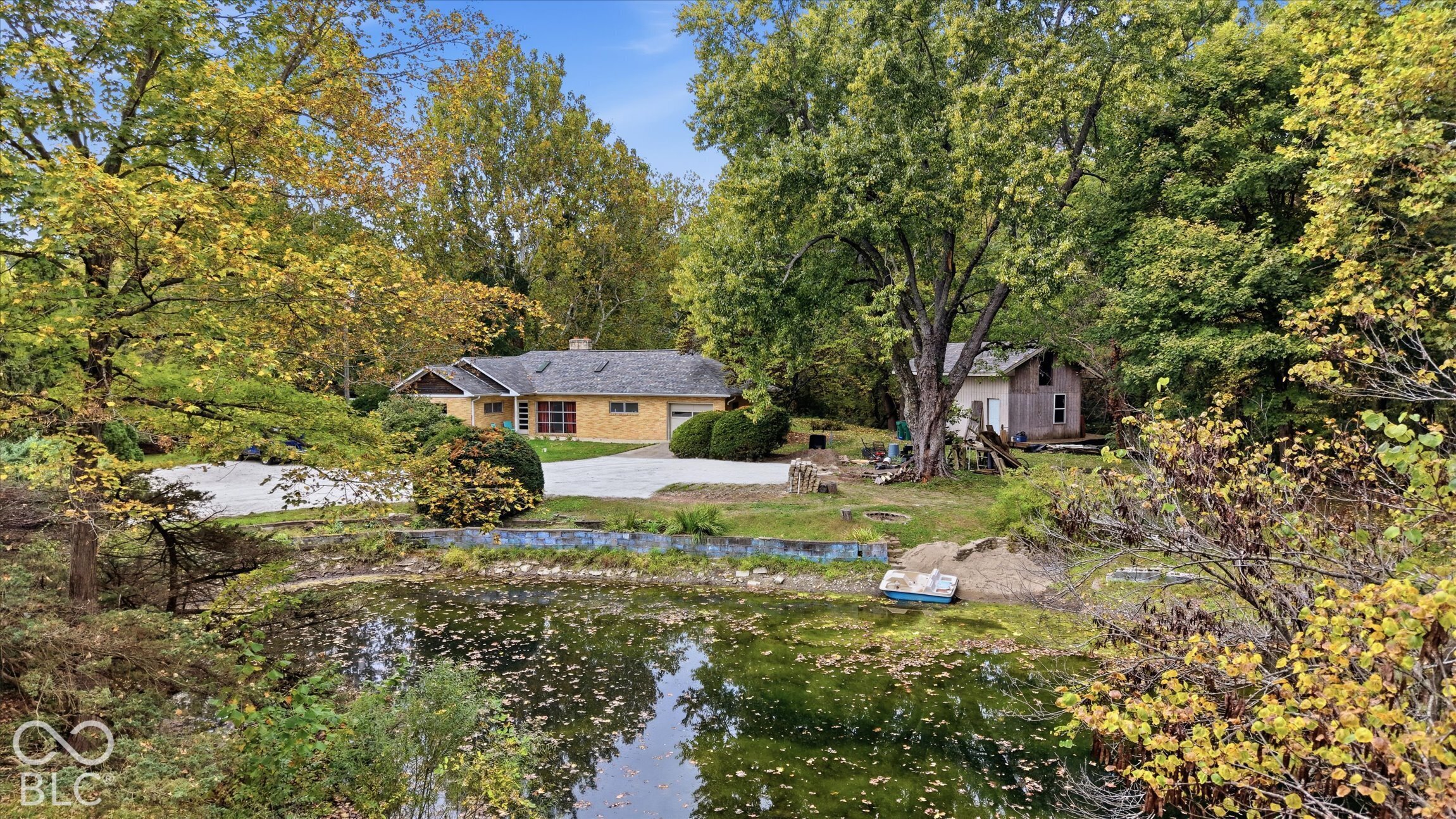 Expansive exterior featuring a natural pond, lush foliage, a secondary structure, a paved driveway, and a brick residence