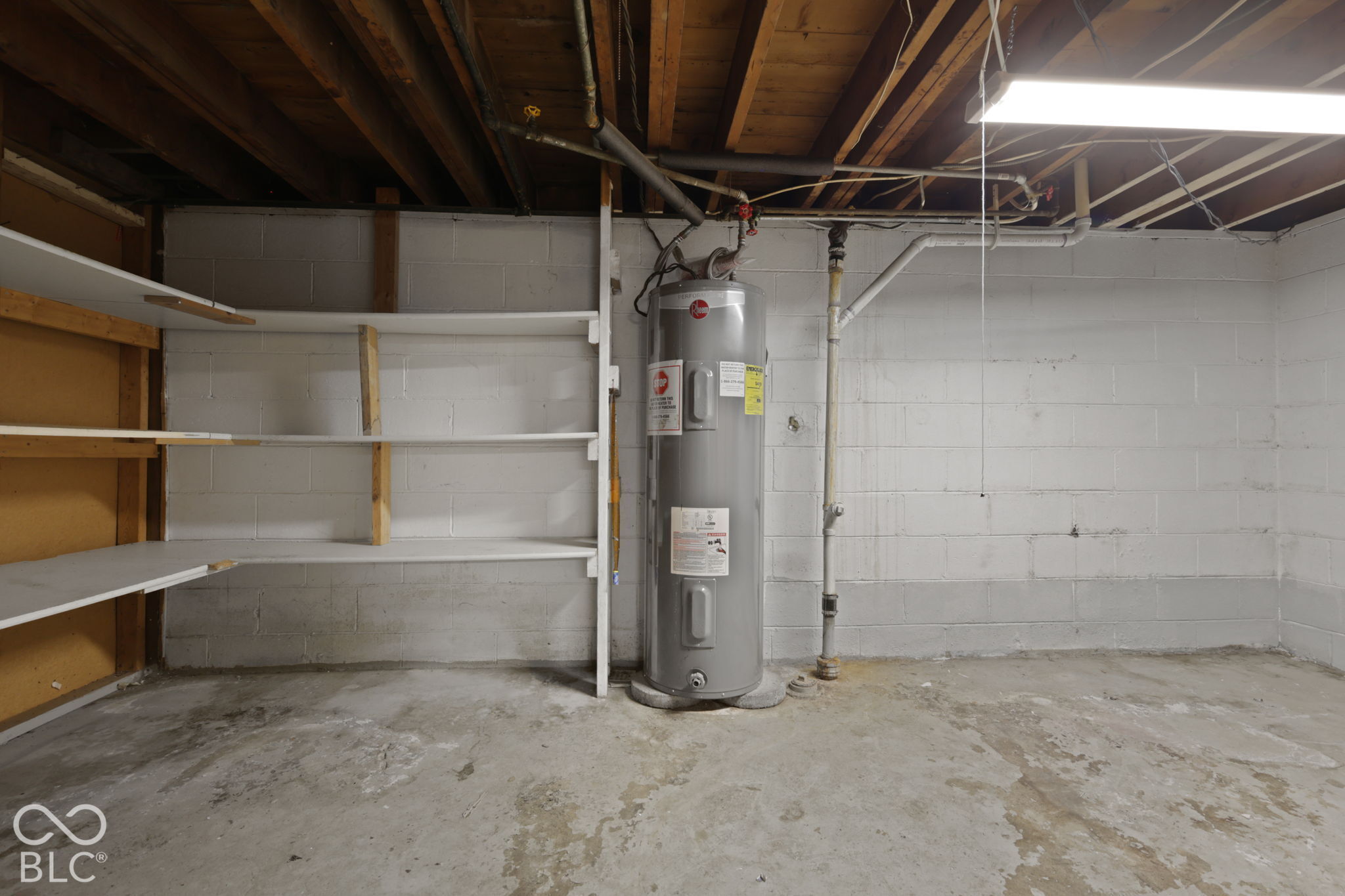 Storage area featuring built-in wooden shelving, painted concrete masonry unit walls, a concrete floor, exposed ceiling joists, and a tall water heater