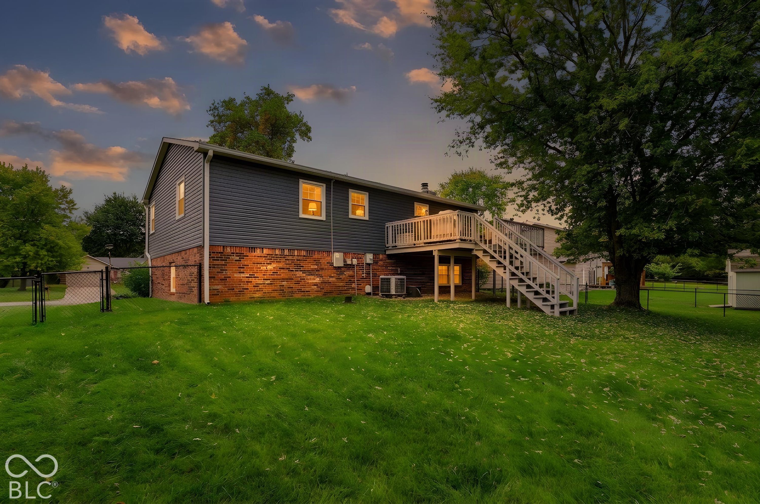 Exterior featuring brick and horizontal siding, elevated wooden deck with stairs, chain-link fencing, and mature trees on a level lawn