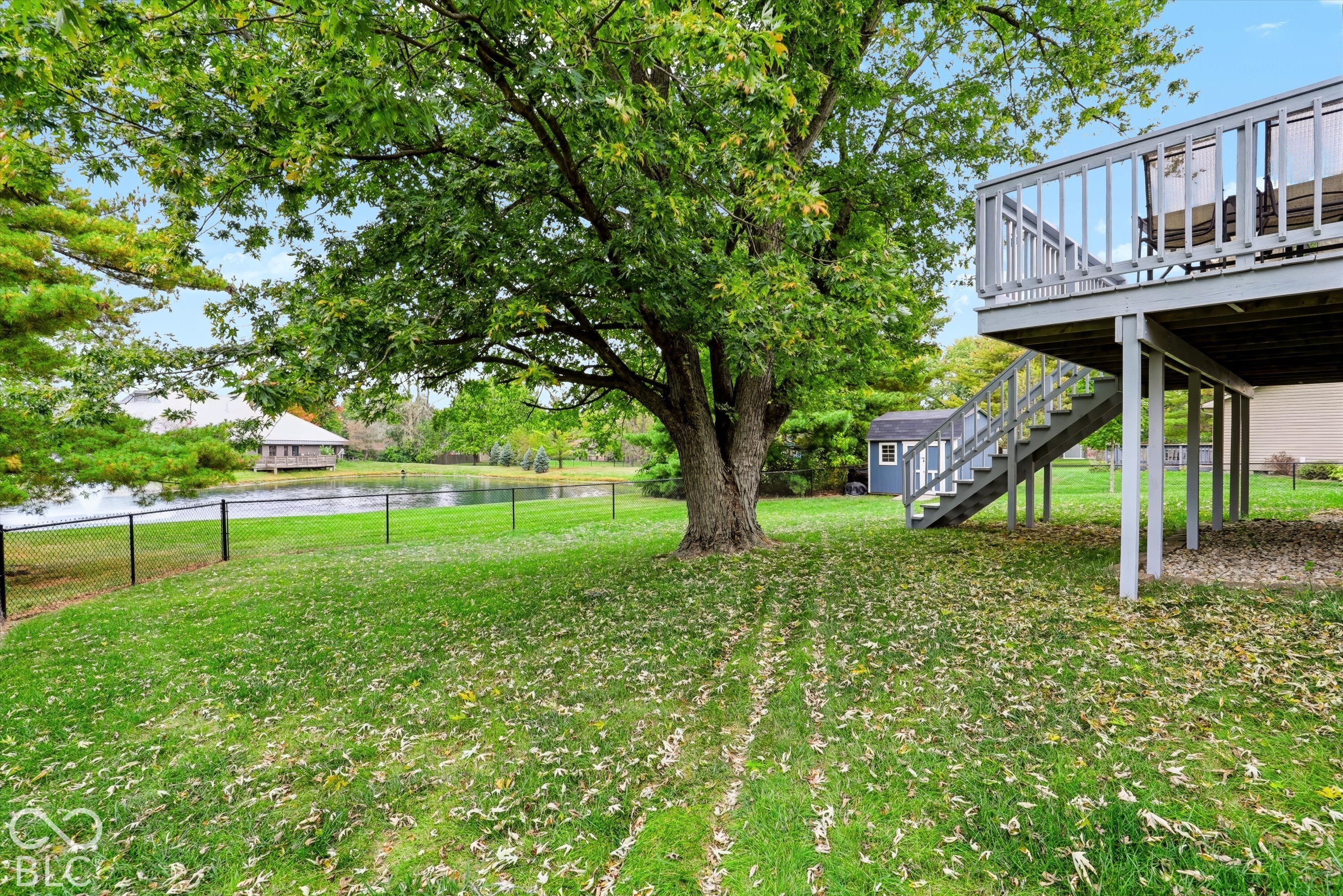 Backyard featuring an elevated wooden deck, staircase, mature tree, storage shed, and chain-link fencing overlooking a pond