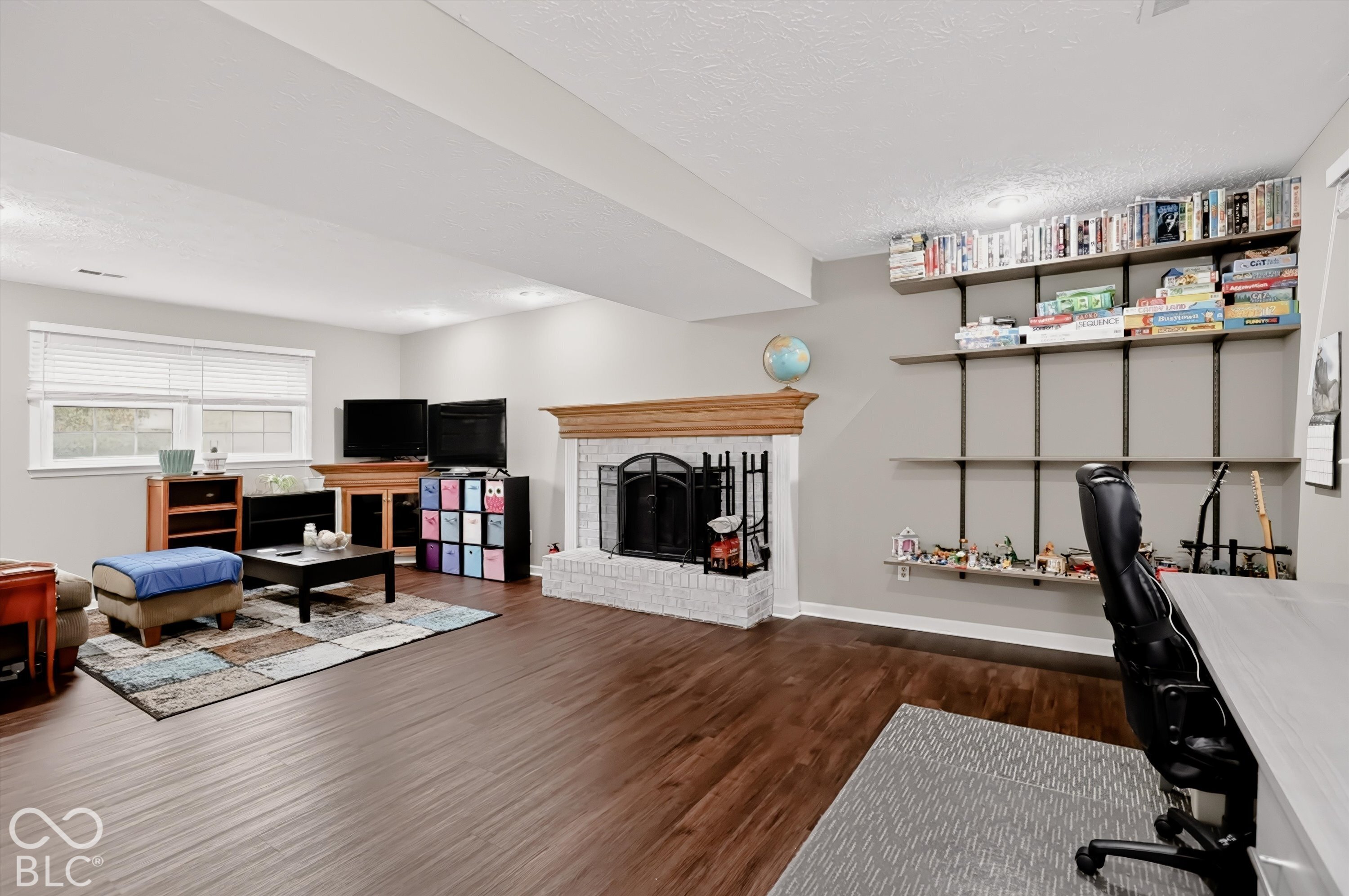 Living area featuring wood-finish flooring, white-painted brick fireplace with a wood mantel, and wall-mounted shelving units