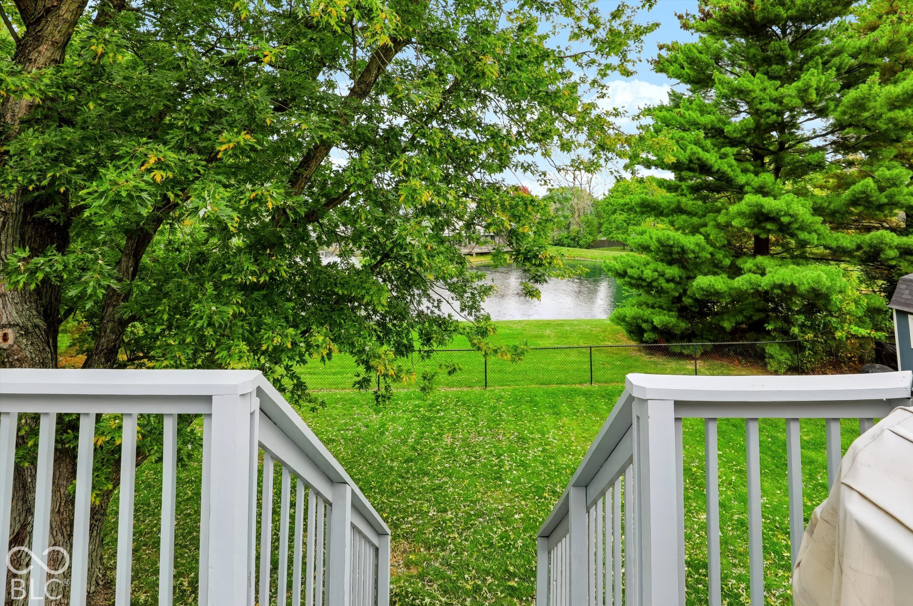 Elevated deck featuring painted railings and staircases overlooking a manicured lawn, mature trees, and a pond
