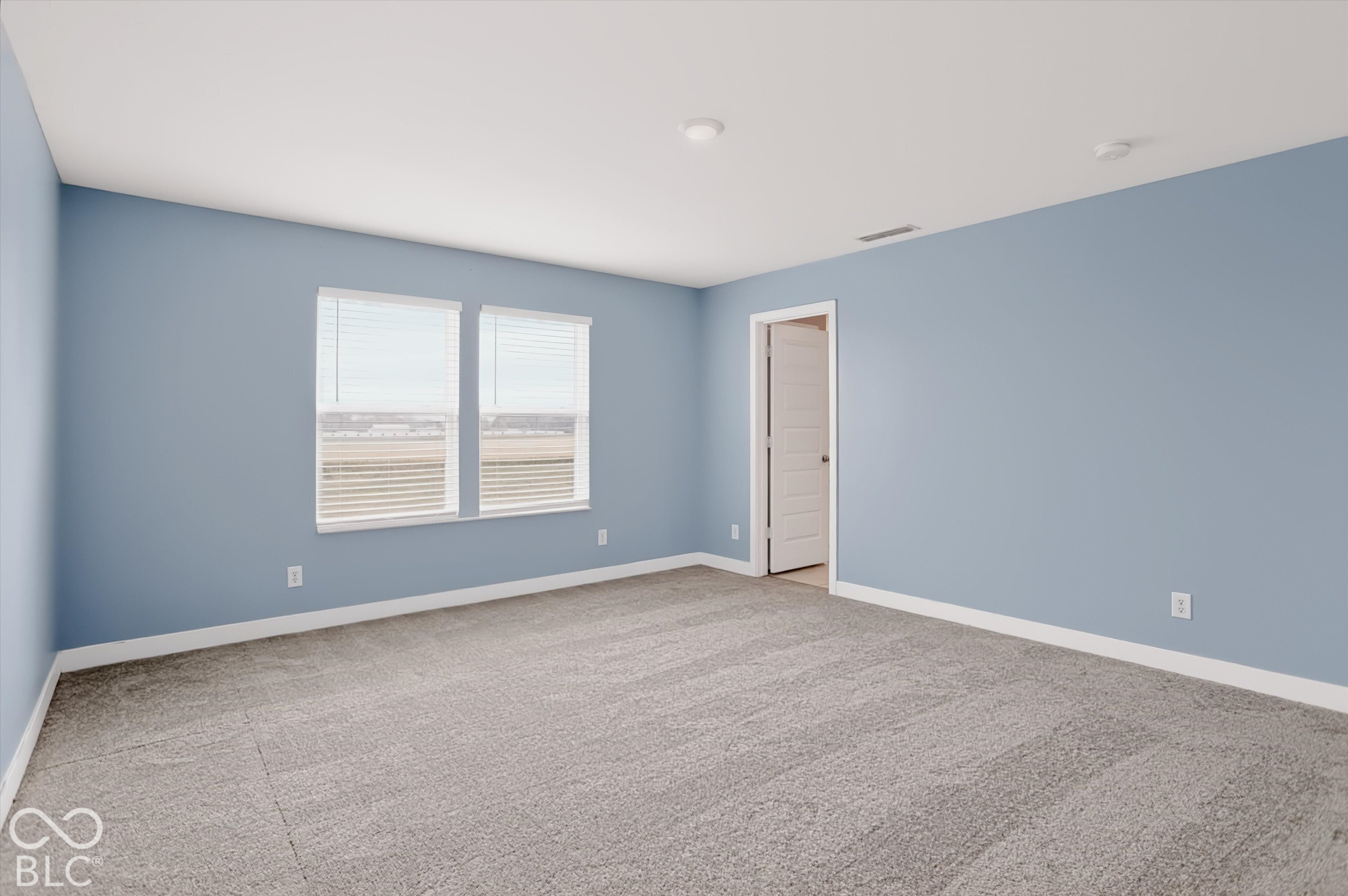 Neutral-toned interior room featuring wall-to-wall carpeting, dual windows with integrated blinds, and a multi-panel interior door