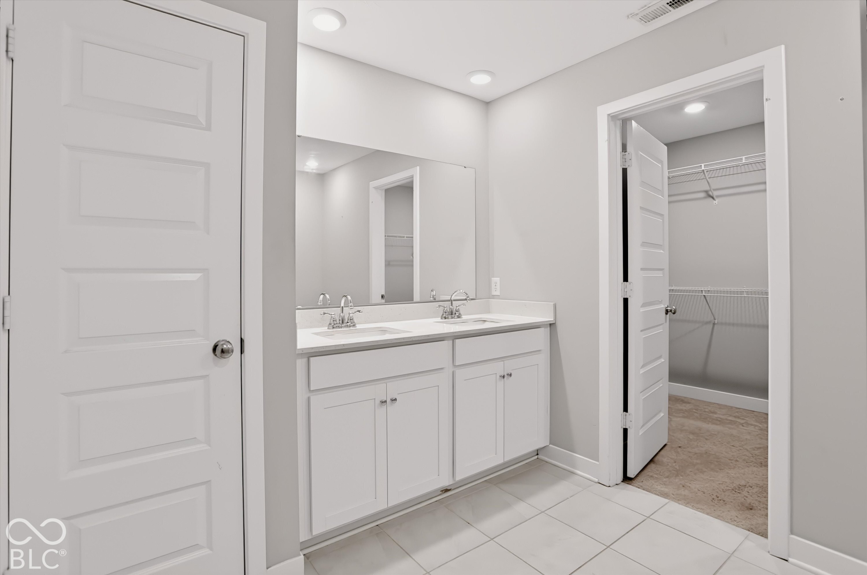 Dual-sink vanity with white cabinetry and solid-surface countertop, full-width wall mirror, recessed lighting, and walk-in closet with wire shelving
