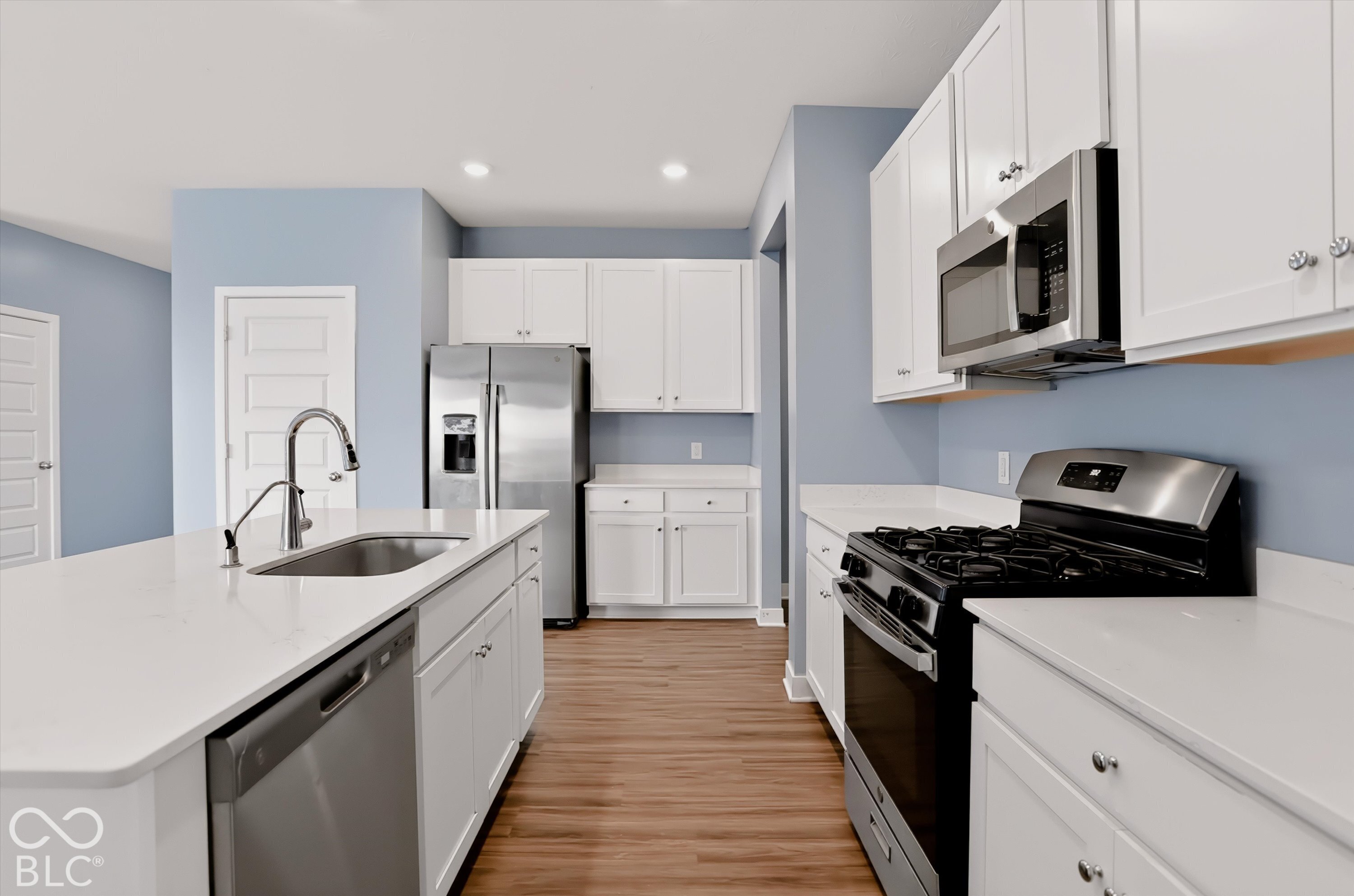 Kitchen featuring white shaker-style cabinetry, a central island with a stainless steel sink, solid-surface countertops, wood-finish flooring, and stainless steel appliances