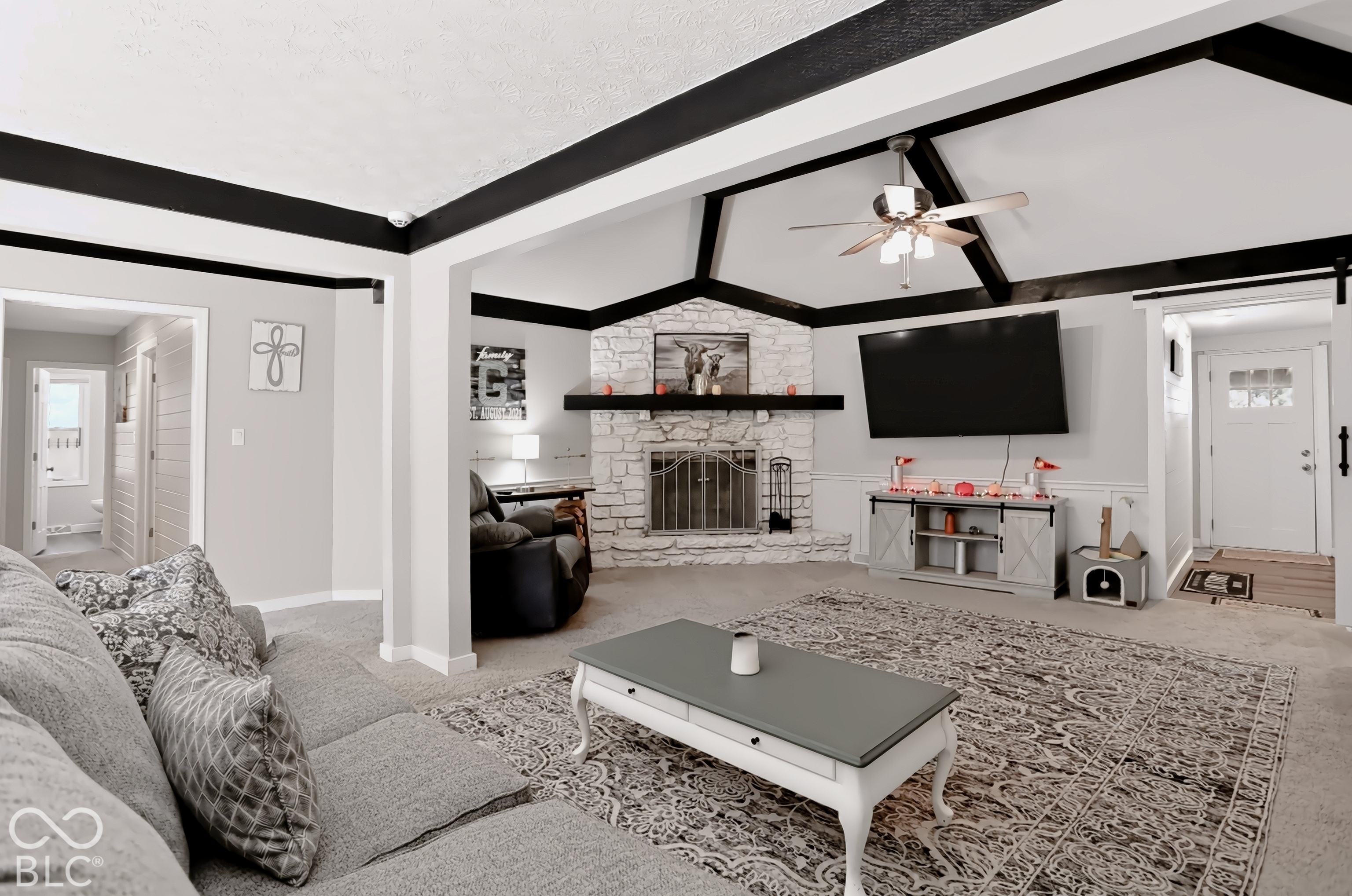 Vaulted ceiling with exposed beams, stone-clad fireplace with mantel, ceiling fan with integrated lighting, paneled accent wall, and a sliding barn door