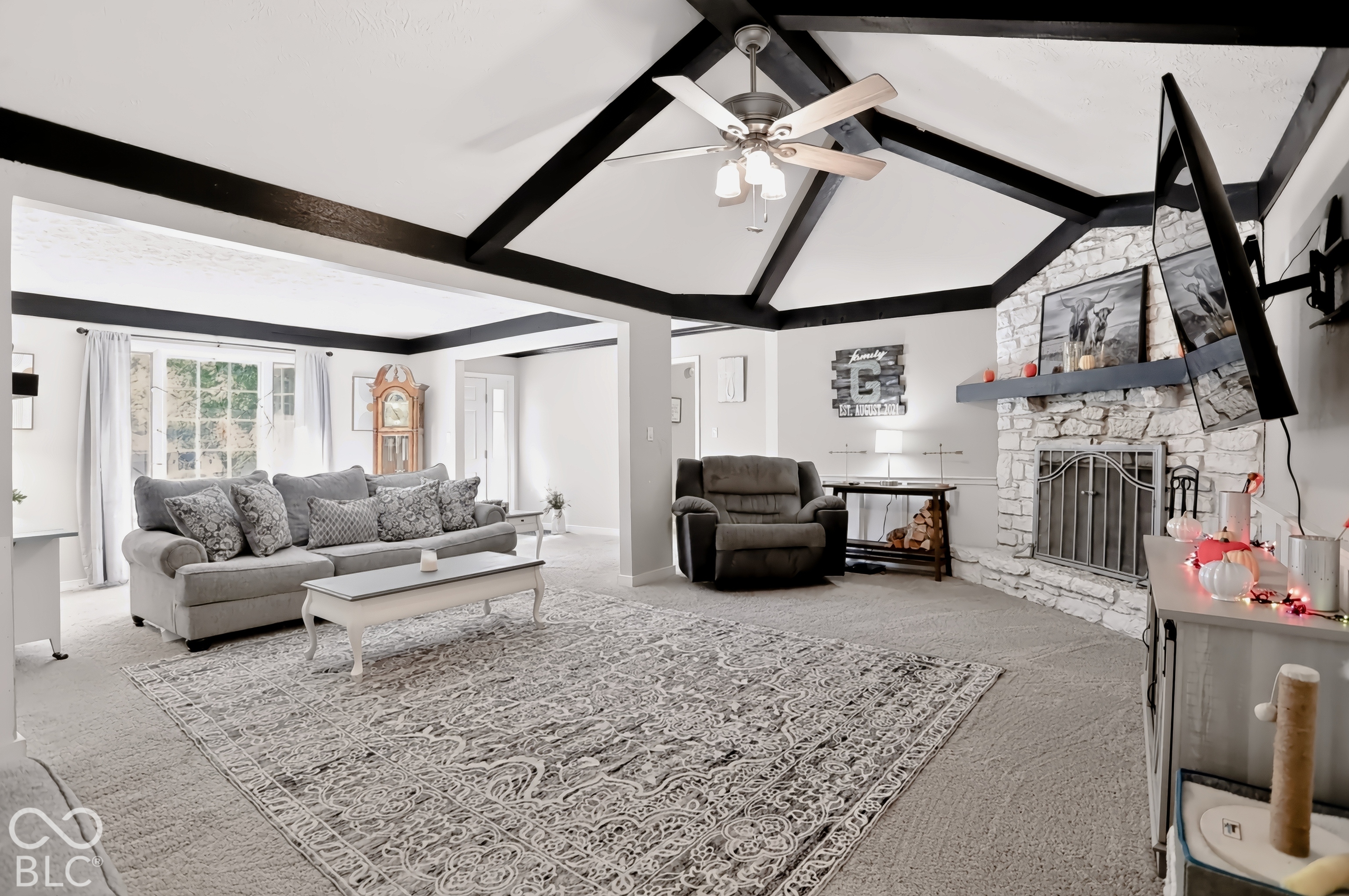 Vaulted ceiling with exposed beams, stone-clad fireplace with mantel, ceiling fan with integrated lighting, built-in wall-mounted television bracket, and large window with white trim